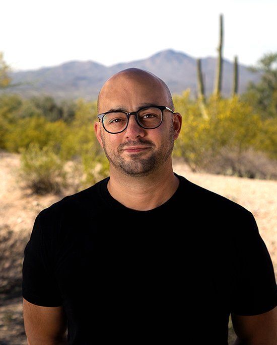 Man with glasses and black shirt standing on a desert trail with mountains and cacti in the background