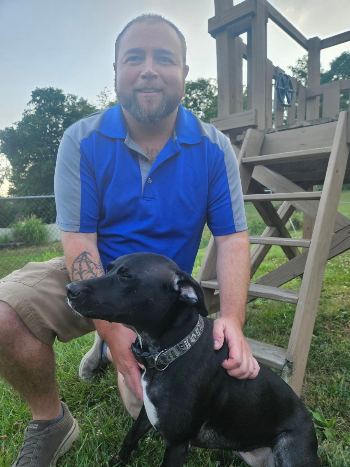 Man in blue shirt crouching beside a black dog outdoors near a wooden play structure