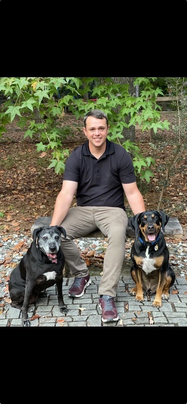 Man seated outdoors with two dogs on a stone path, greenery and brick wall behind them.