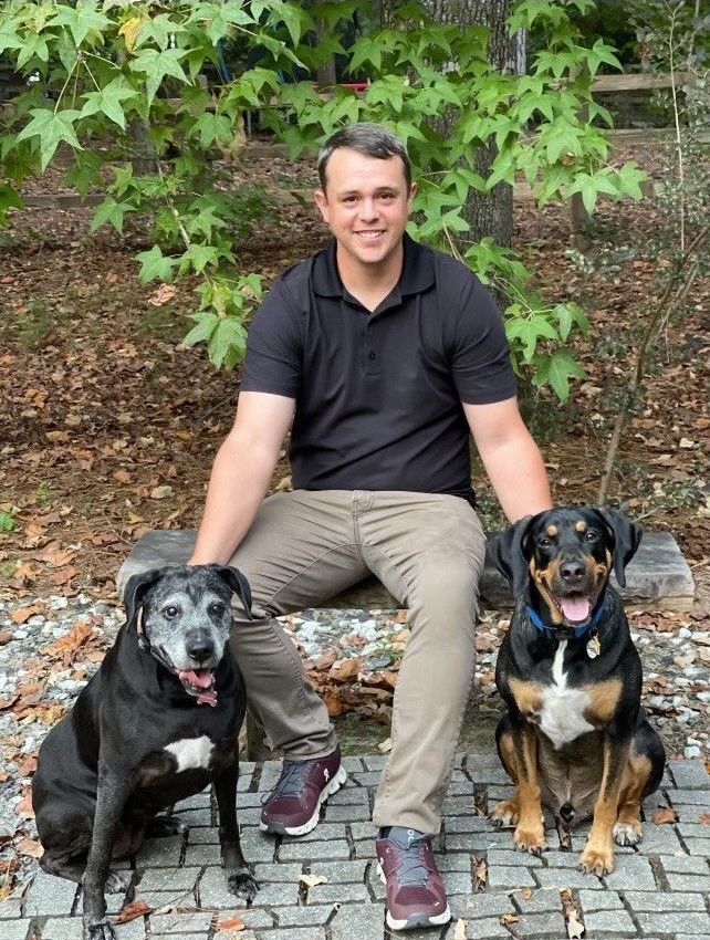 Man sitting on a stone bench with two dogs in a leafy outdoor setting