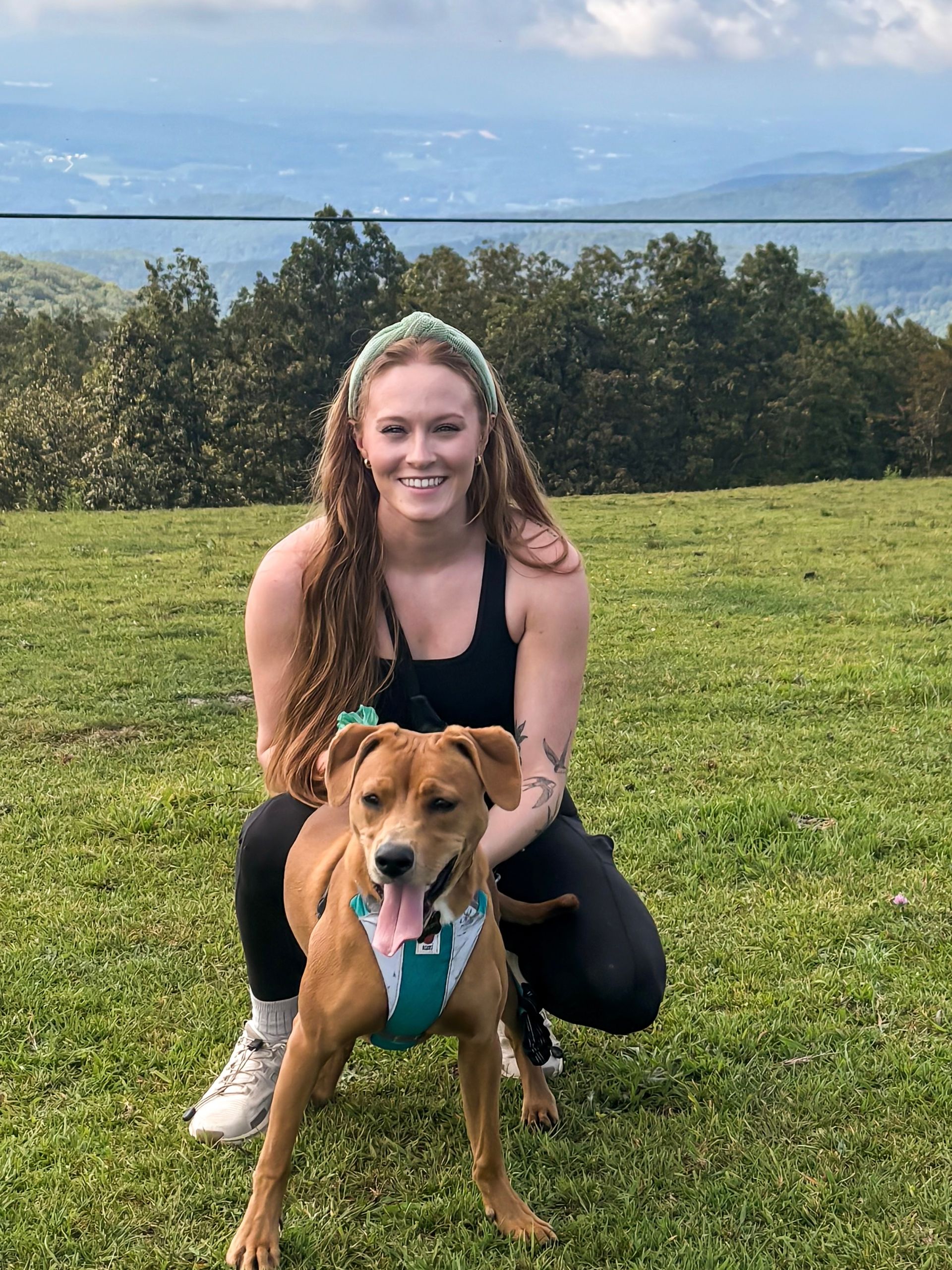 Woman crouching behind a brown dog on a grassy hill, with mountains and trees in the background.