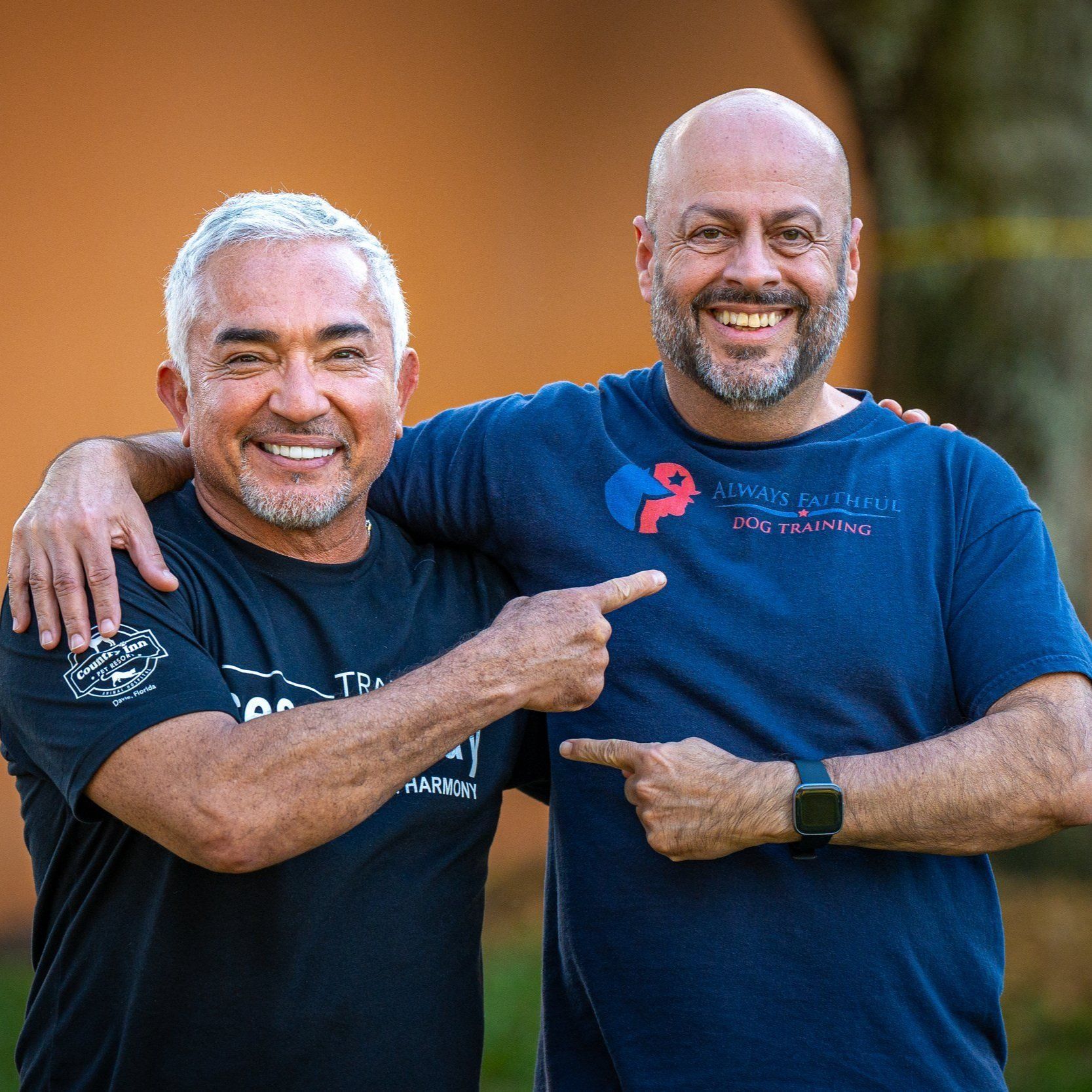 Two smiling men in navy shirts pointing at each other outdoors