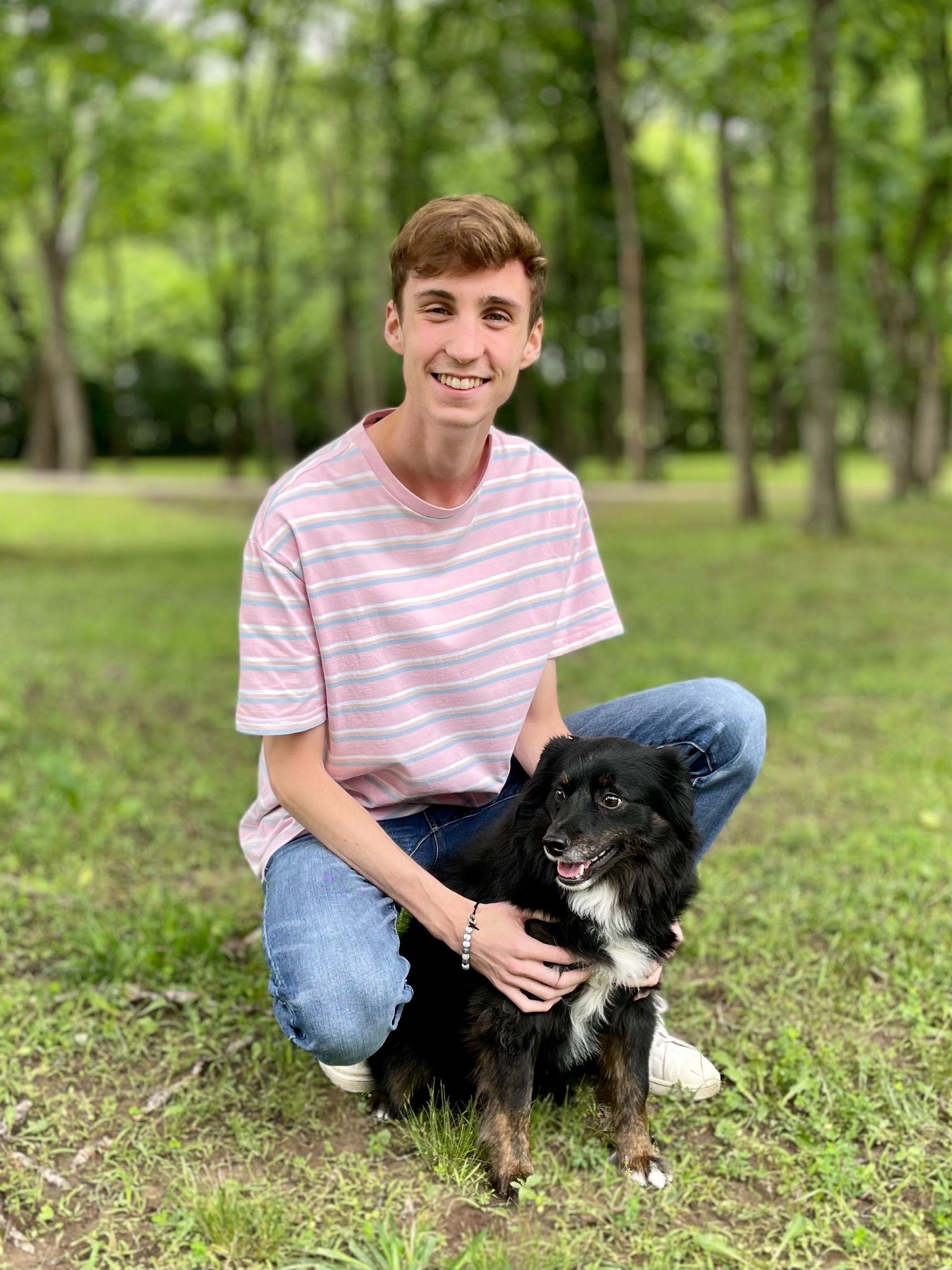Young man kneeling on grass with a small black dog in a leafy park