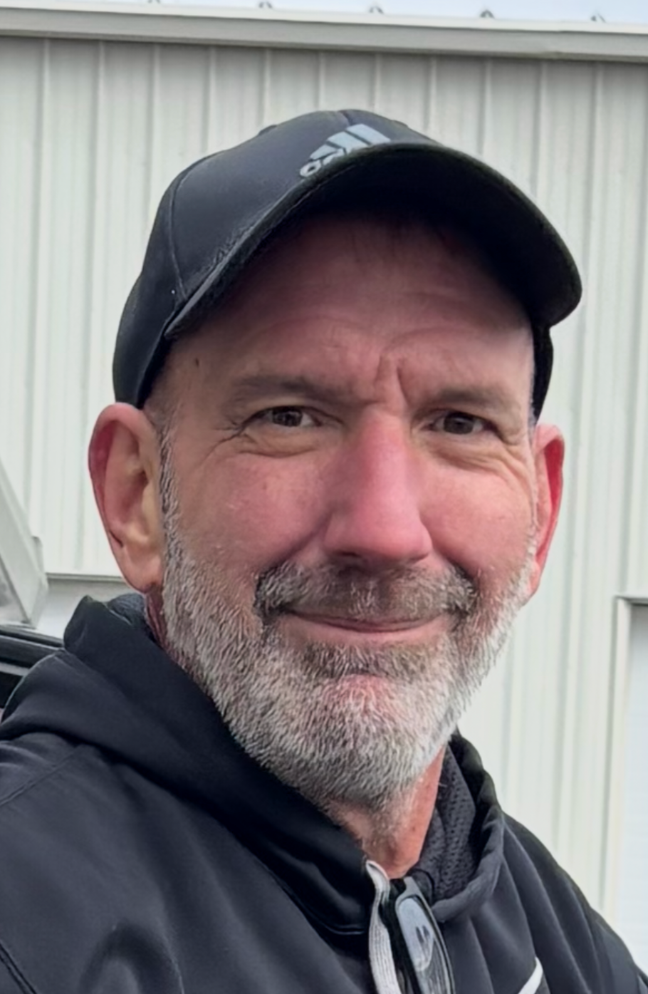 Man in black cap and jacket smiling outdoors in front of a light gray building.