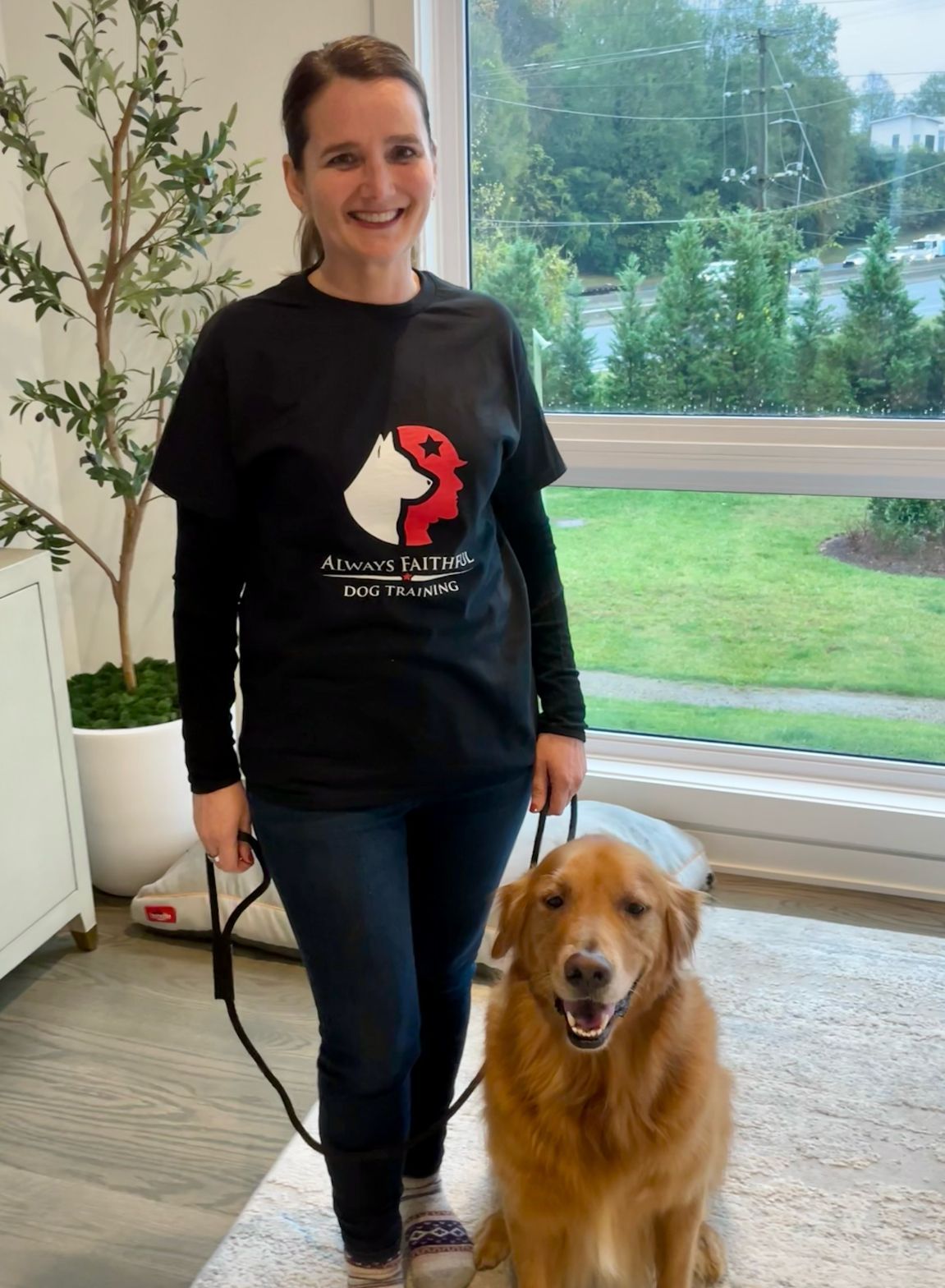 Woman in black shirt standing indoors beside a golden retriever by a window with greenery outside