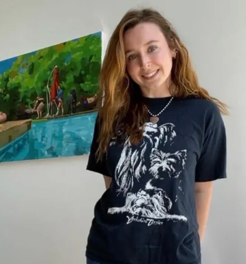 Woman smiling indoors beside a colorful pool painting, wearing a black graphic T-shirt.