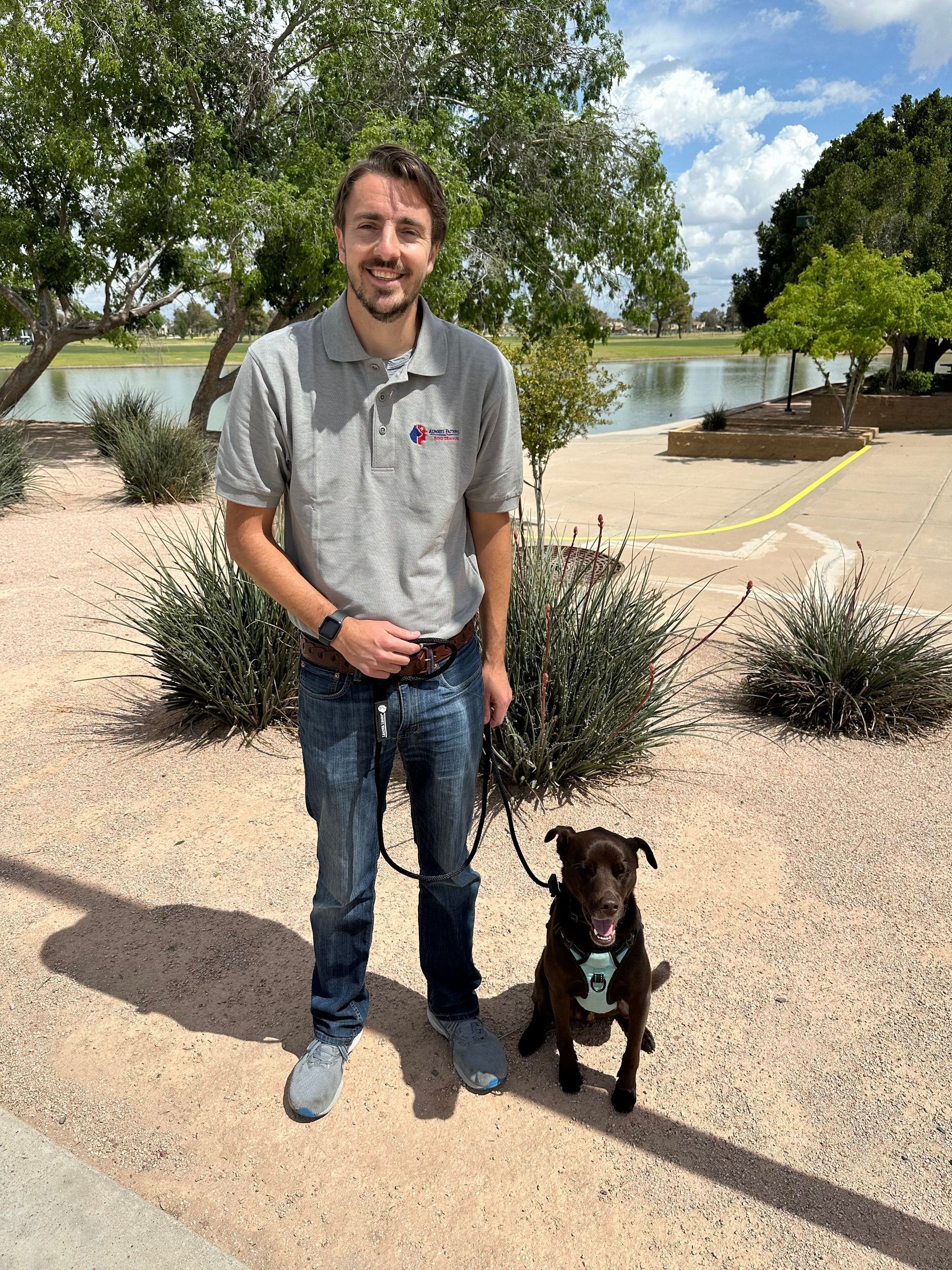 Man standing with a small dog beside him in a sunny park near a lake