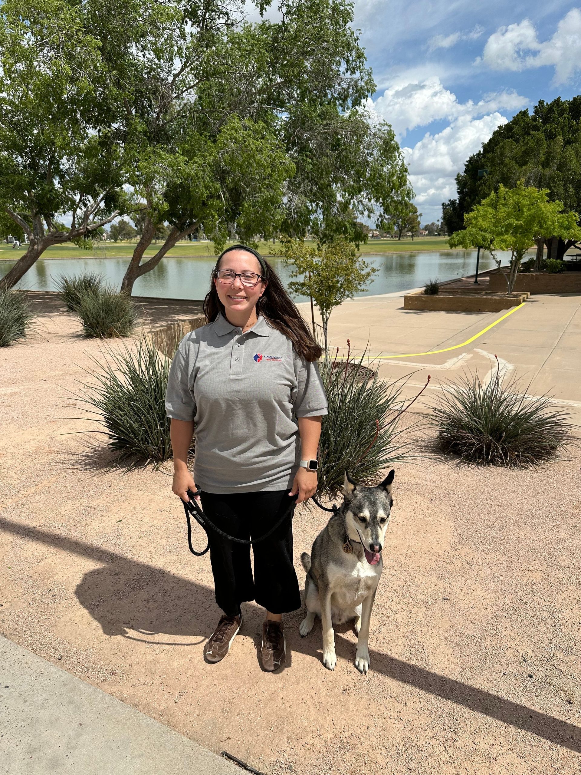 Person standing with a dog on a lakeside path, with trees, rocks, and water in the background.