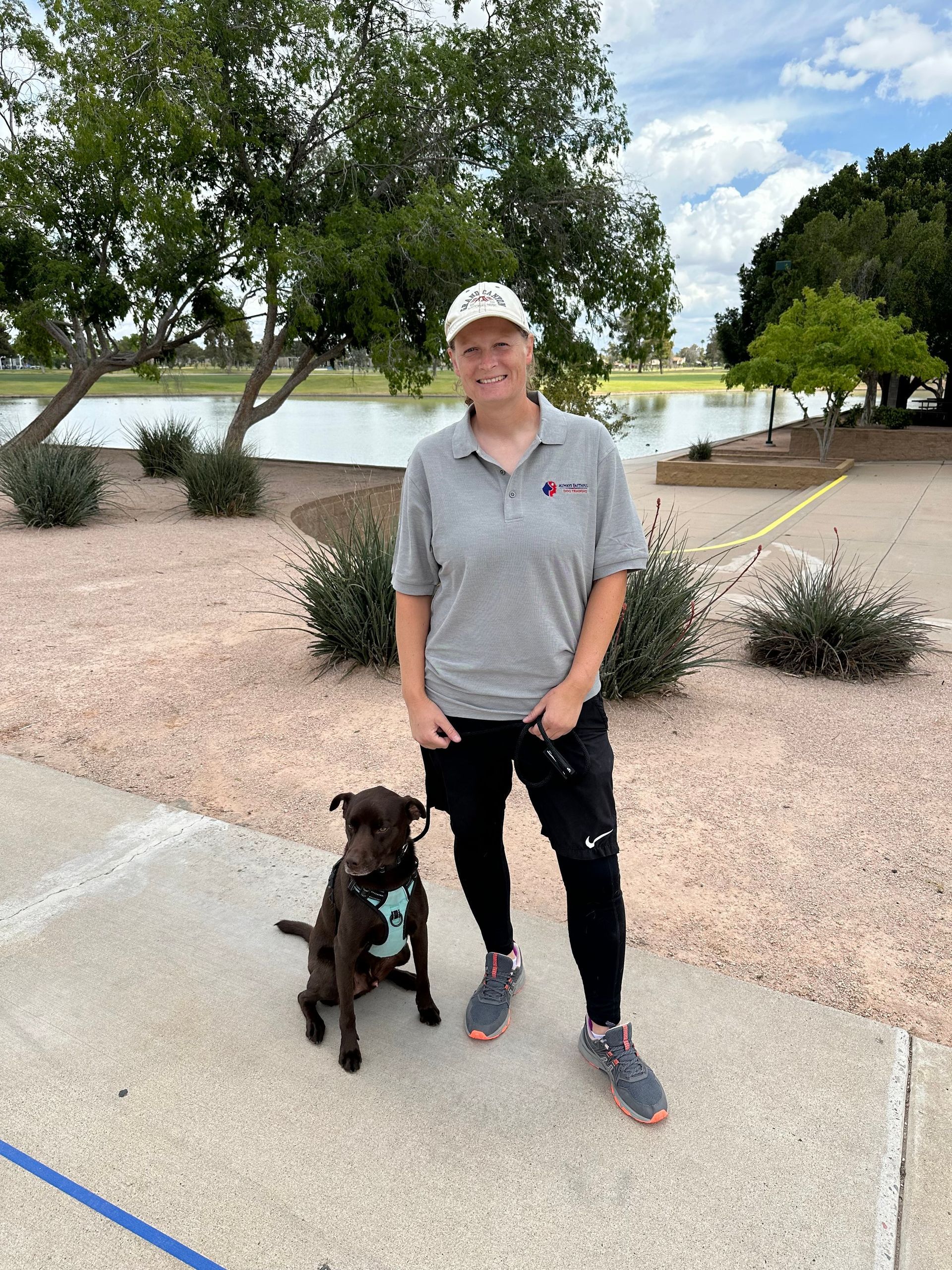 Person standing with a black dog on a sidewalk beside trees and a lake