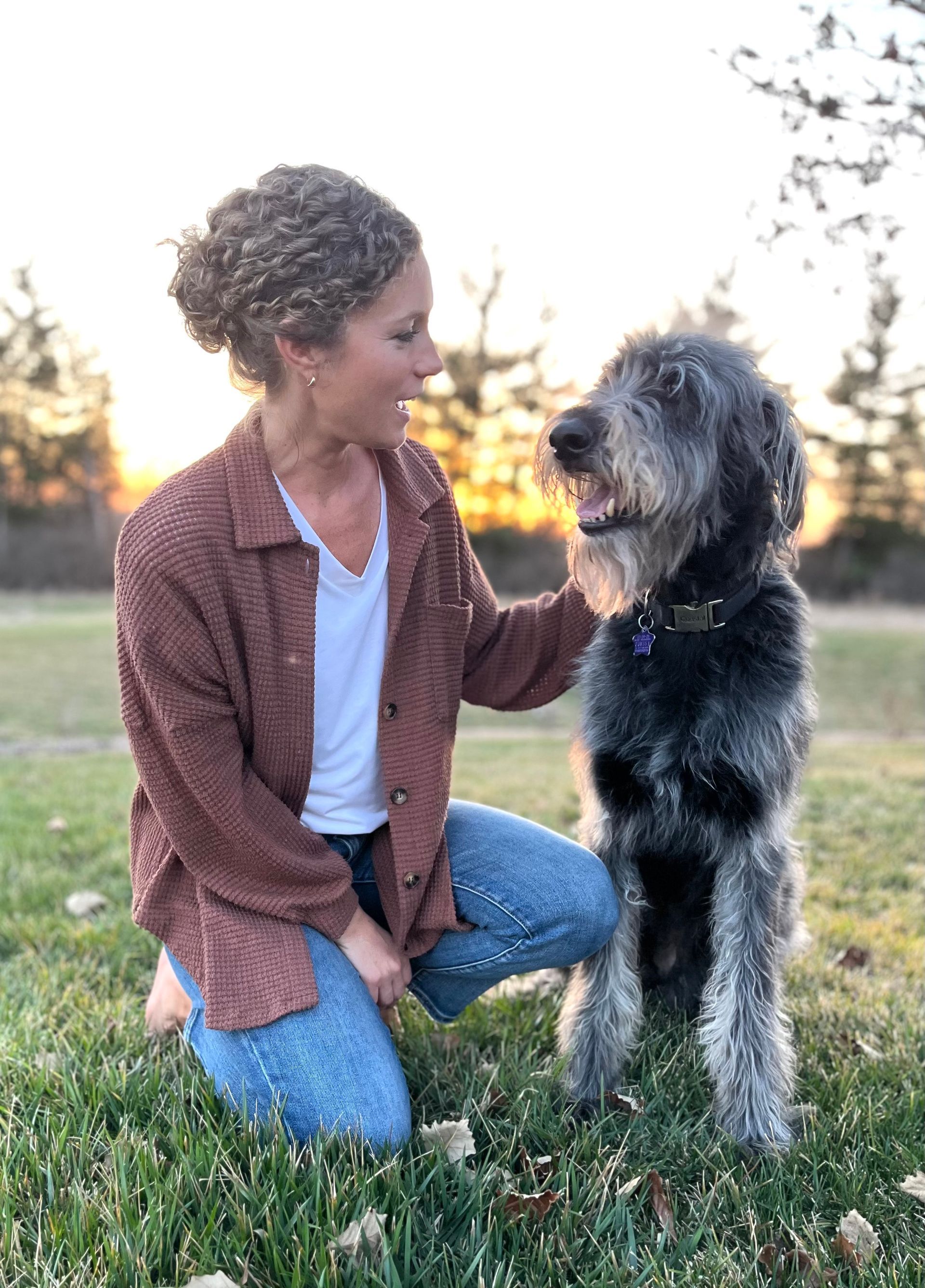 Woman kneels petting a shaggy black dog in a grassy field at sunset.