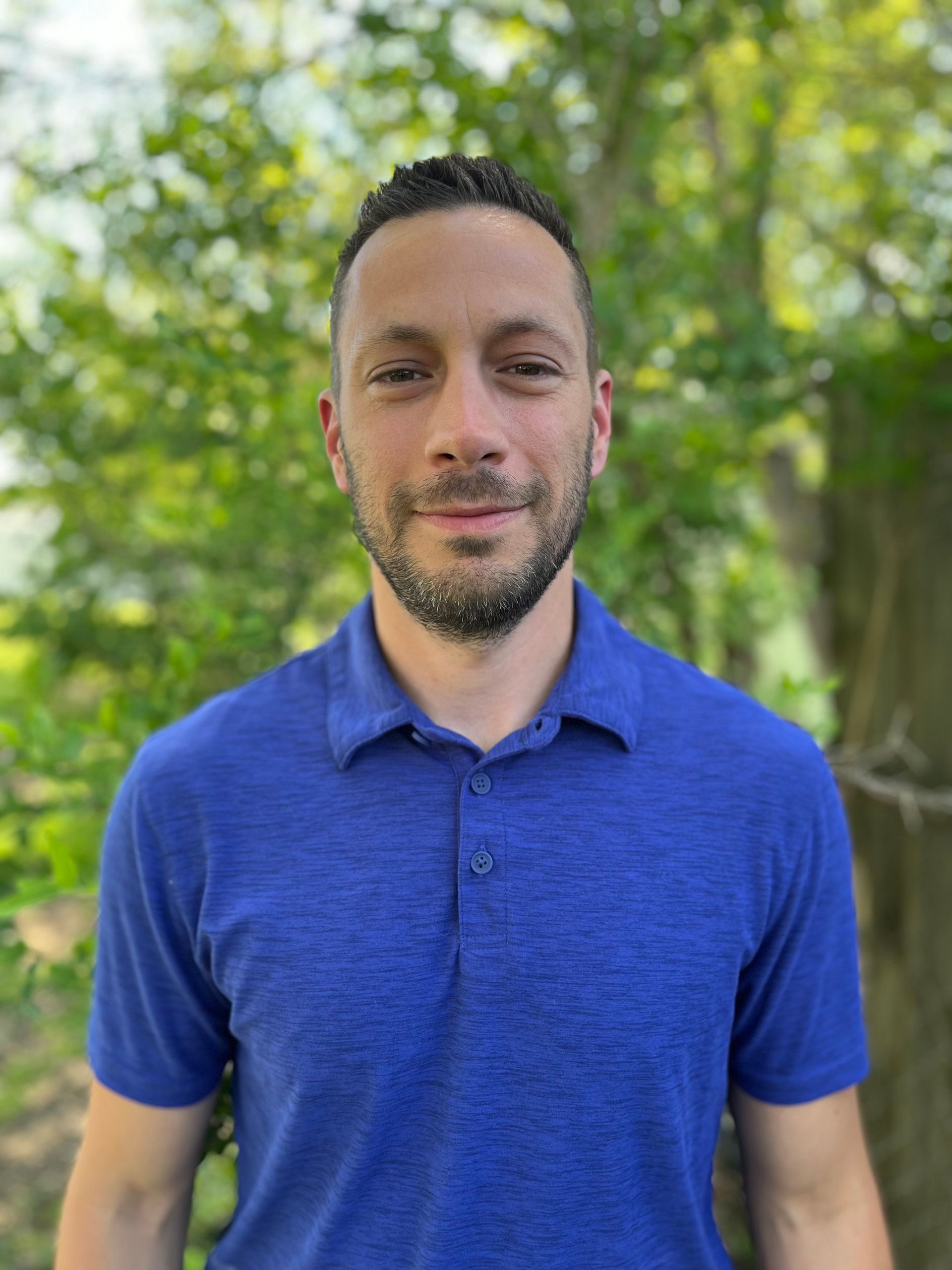 Man in a blue polo shirt standing outdoors in front of green trees, looking at the camera.