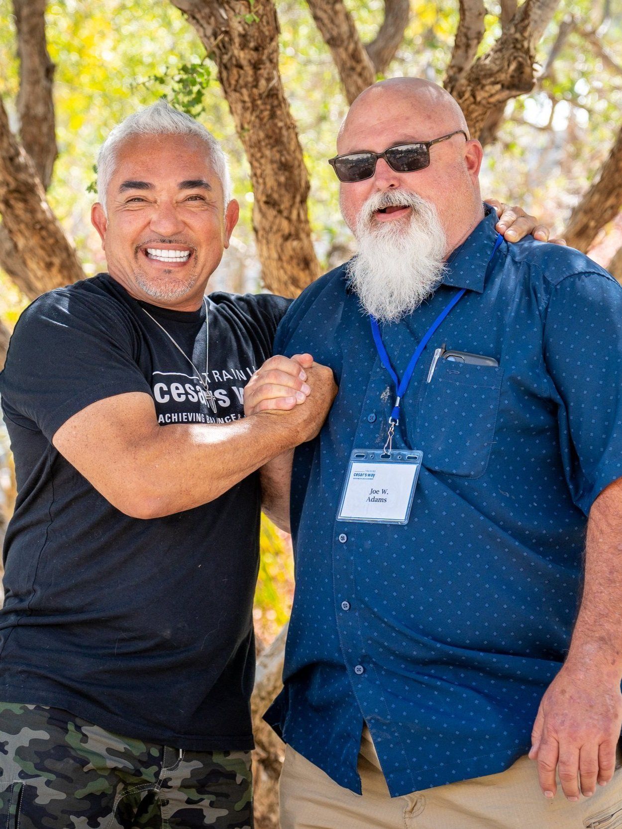 Two smiling men standing outdoors under trees, posing arm-in-arm at an event.