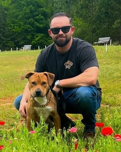 Man kneeling with a brown dog in a grassy field with red flowers and trees in the background