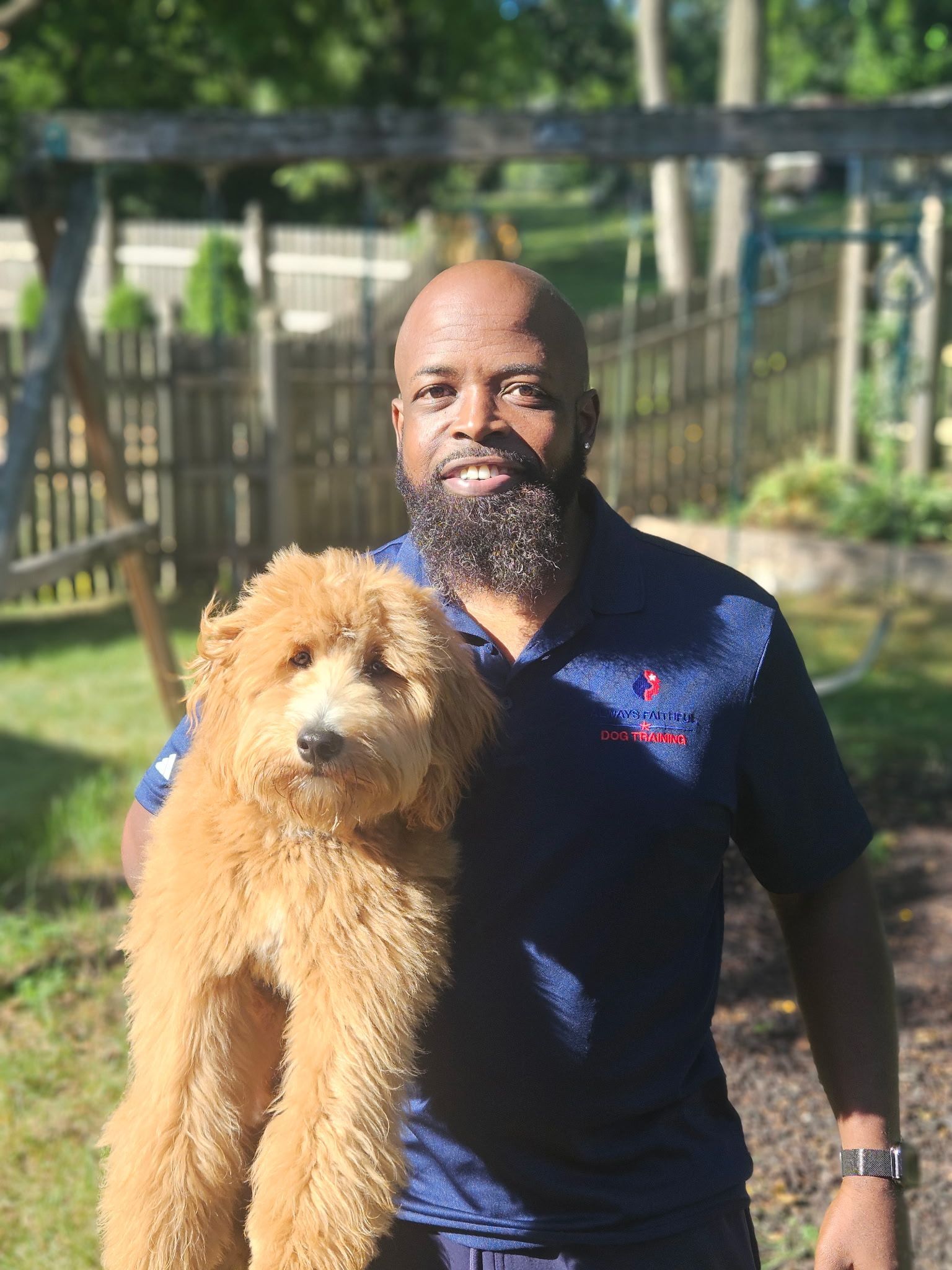 Man in navy shirt holding a fluffy tan dog outdoors near a fence and greenery.
