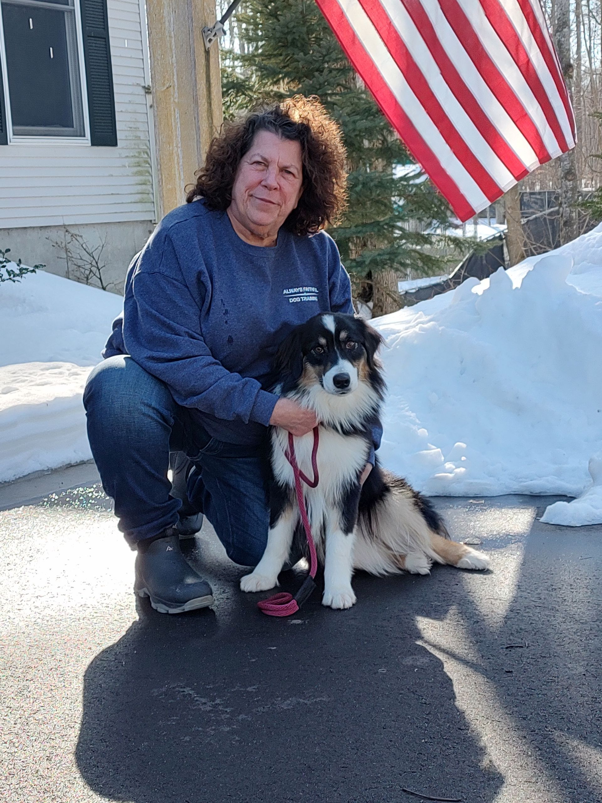 Person kneels beside a black-and-white dog in the snow, with an American flag and house in the background