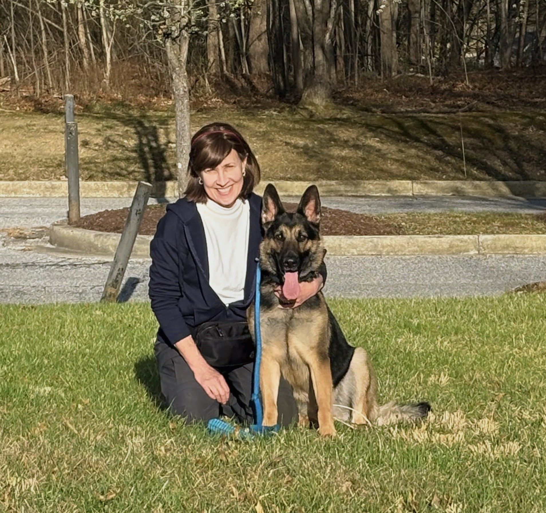 Woman kneeling with German shepherd on grass, both facing camera, on a sunny path near trees