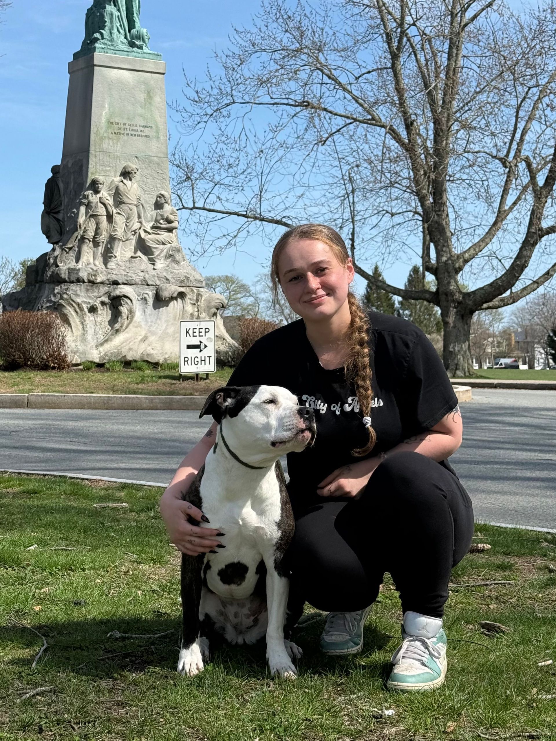 Woman crouches with black-and-white dog on grass near a monument under a clear blue sky.