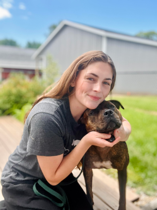 Young woman crouches outdoors, hugging a brown dog beside a sidewalk and grassy yard.