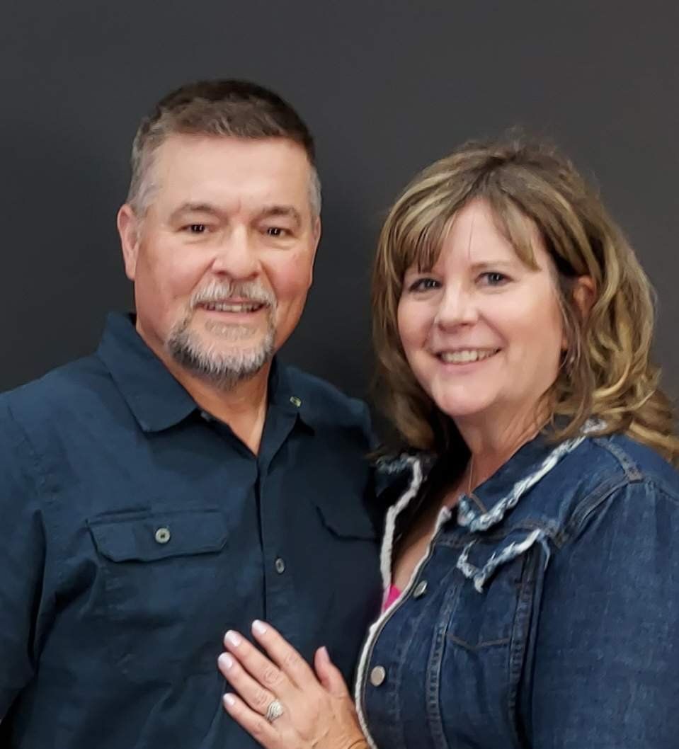 Smiling couple posing together in dark denim jackets against a dark background