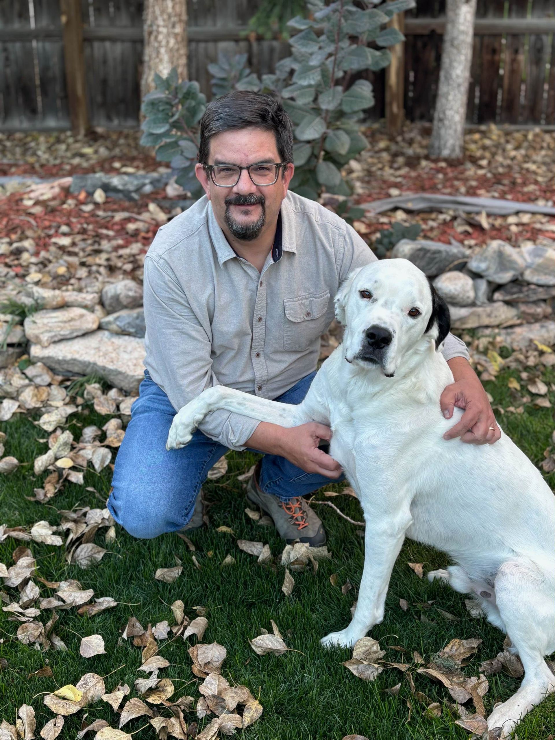 Man kneeling on grass beside a white dog in a backyard with fallen leaves and a stone border
