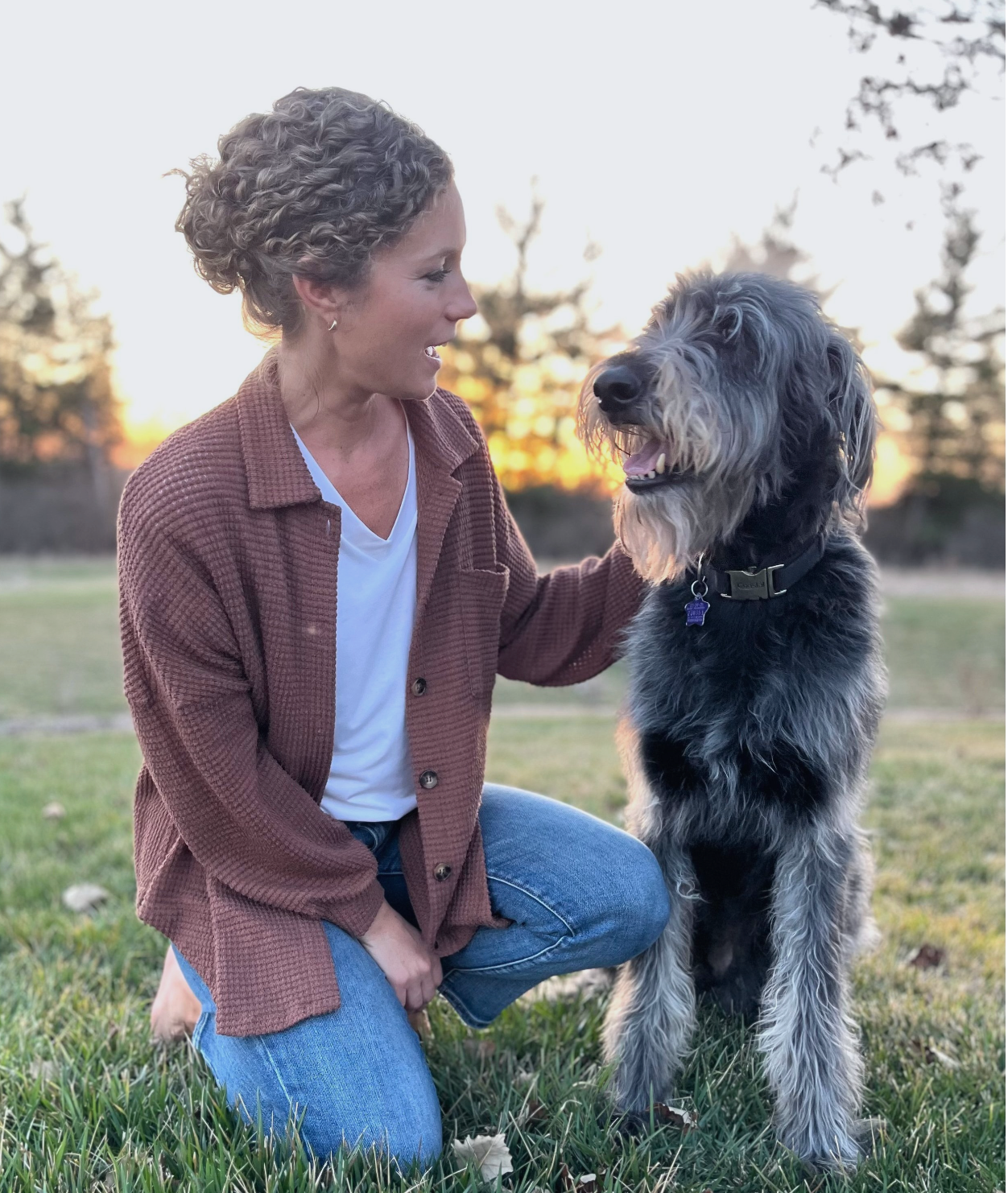 Woman kneeling on grass beside a large shaggy dog at sunset, smiling and petting it.