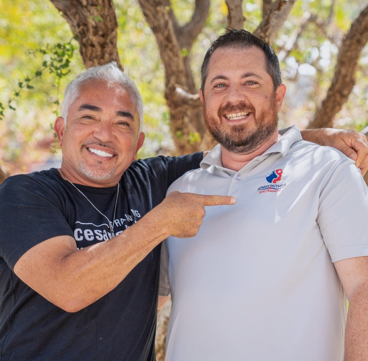 Two smiling men standing outdoors, one pointing at the other, with trees in the background