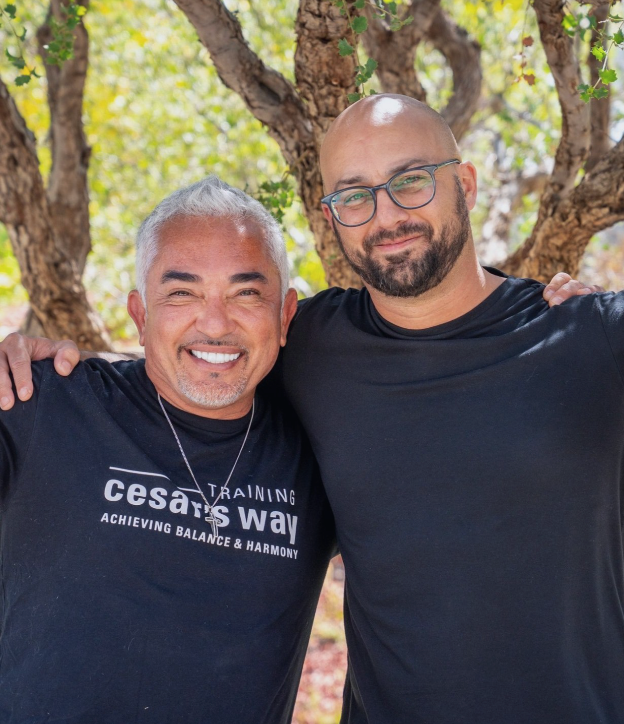 Two smiling men standing arm-in-arm outdoors in front of a tree.