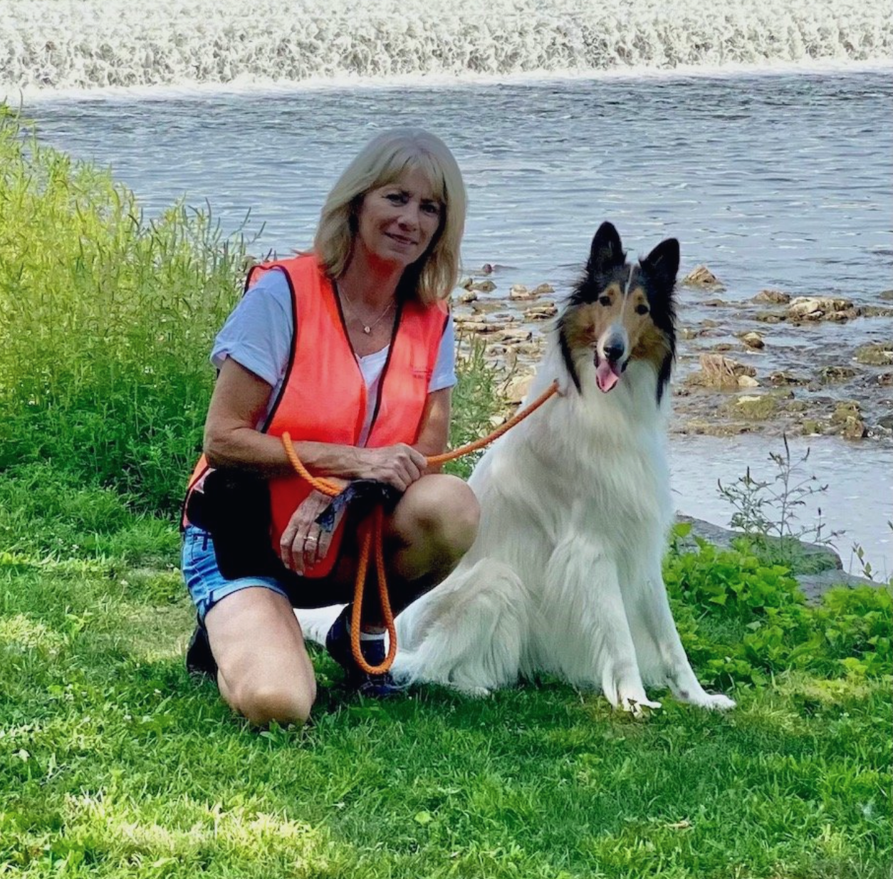 Woman kneeling with a collie on grass by a lakeside, both facing the camera.