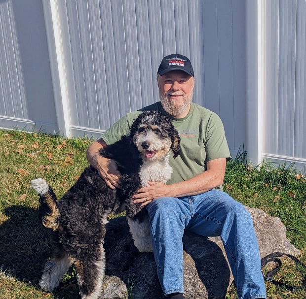 Man in a cap sitting on a rock, smiling with a black-and-white dog beside him near a metal fence.