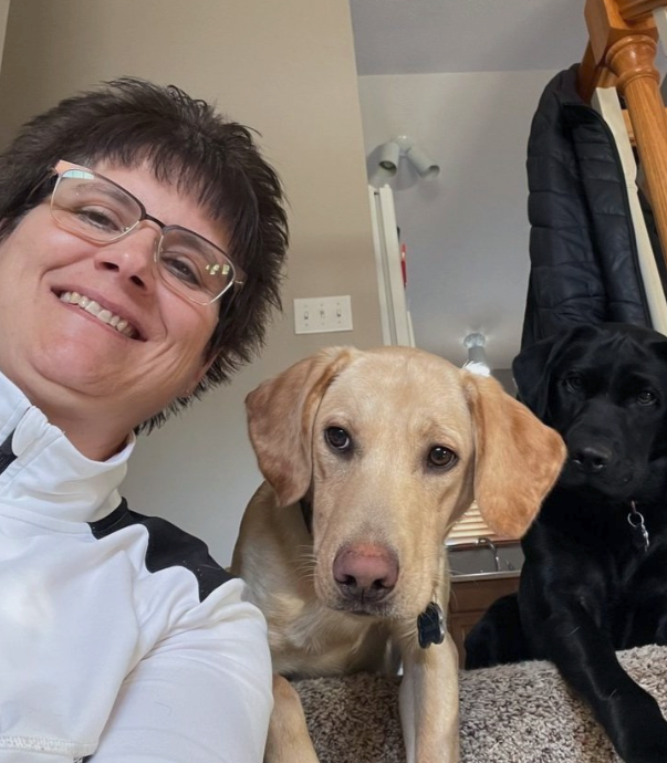 Smiling person with two dogs indoors, including a tan Labrador in the foreground and a black dog behind.