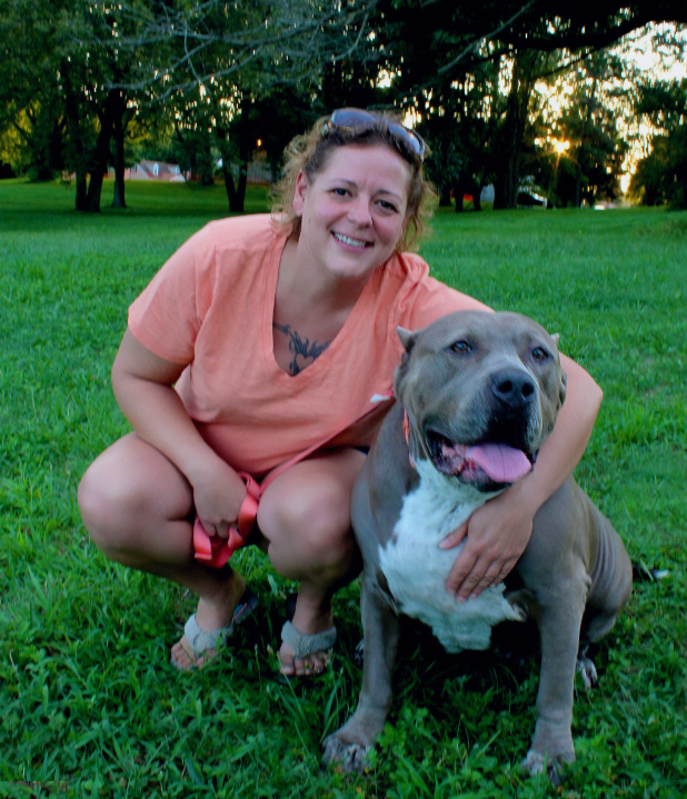 Woman crouching on grass with a gray dog, both smiling in a park at sunset