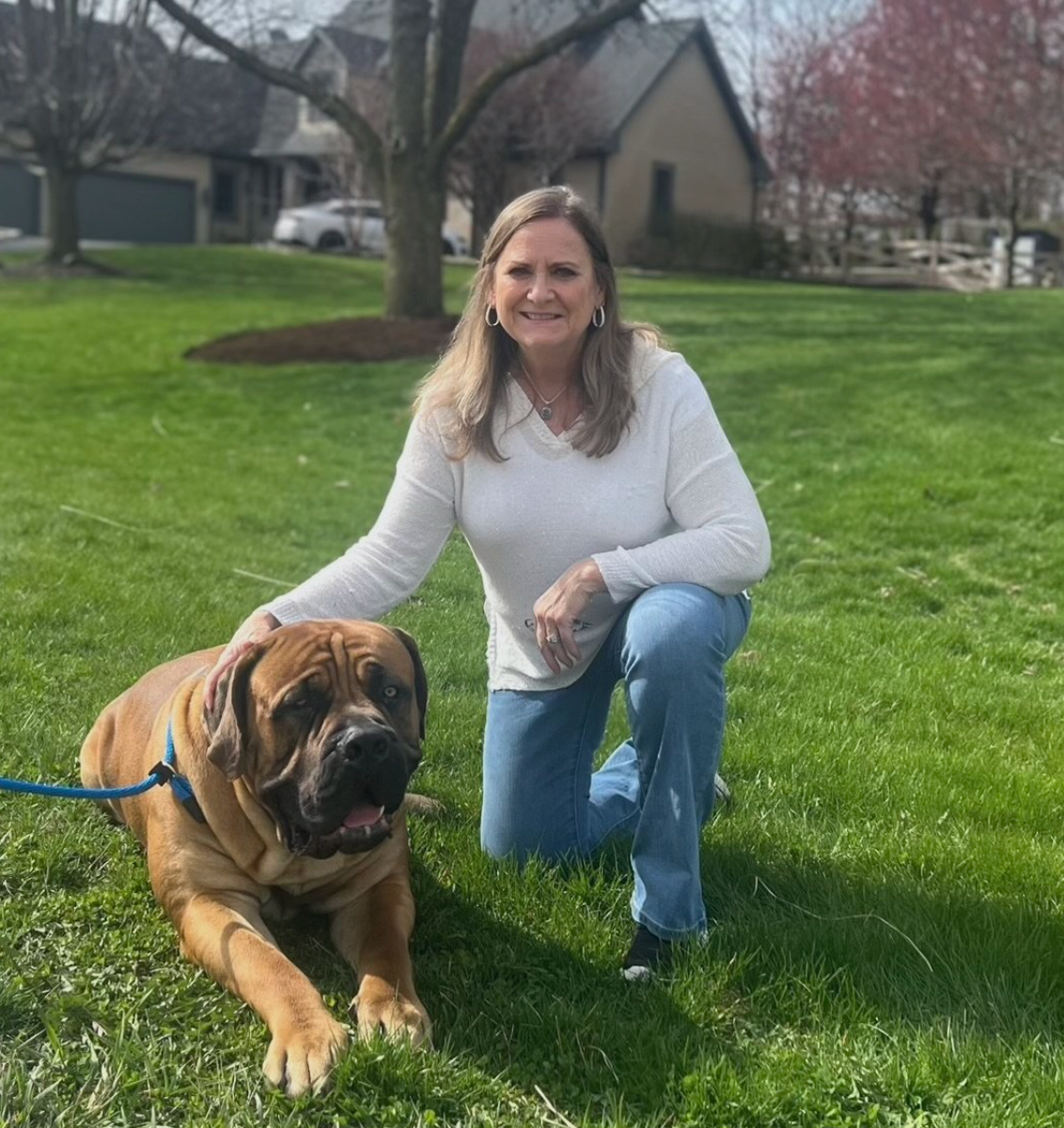 Woman kneeling on grass beside a large brown dog on a leash in a sunny park