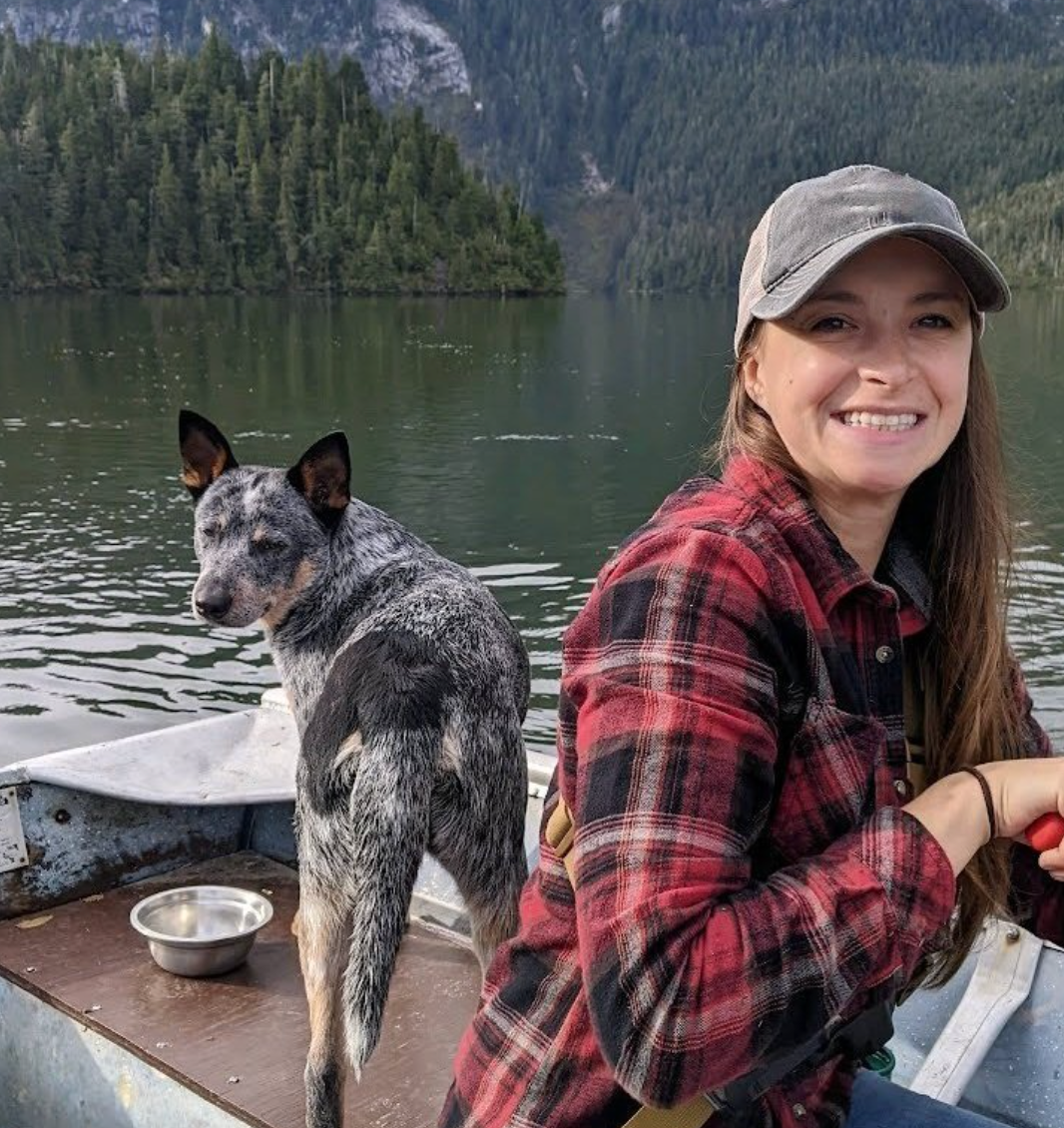 Woman in a red plaid shirt smiles on a boat with a speckled dog on a mountain lake
