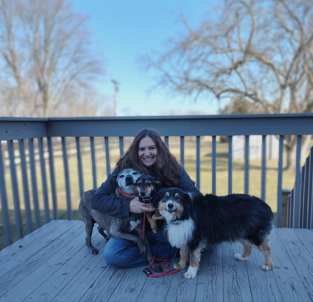 Woman kneeling with two dogs on a wooden deck, smiling outdoors by bare trees under a blue sky