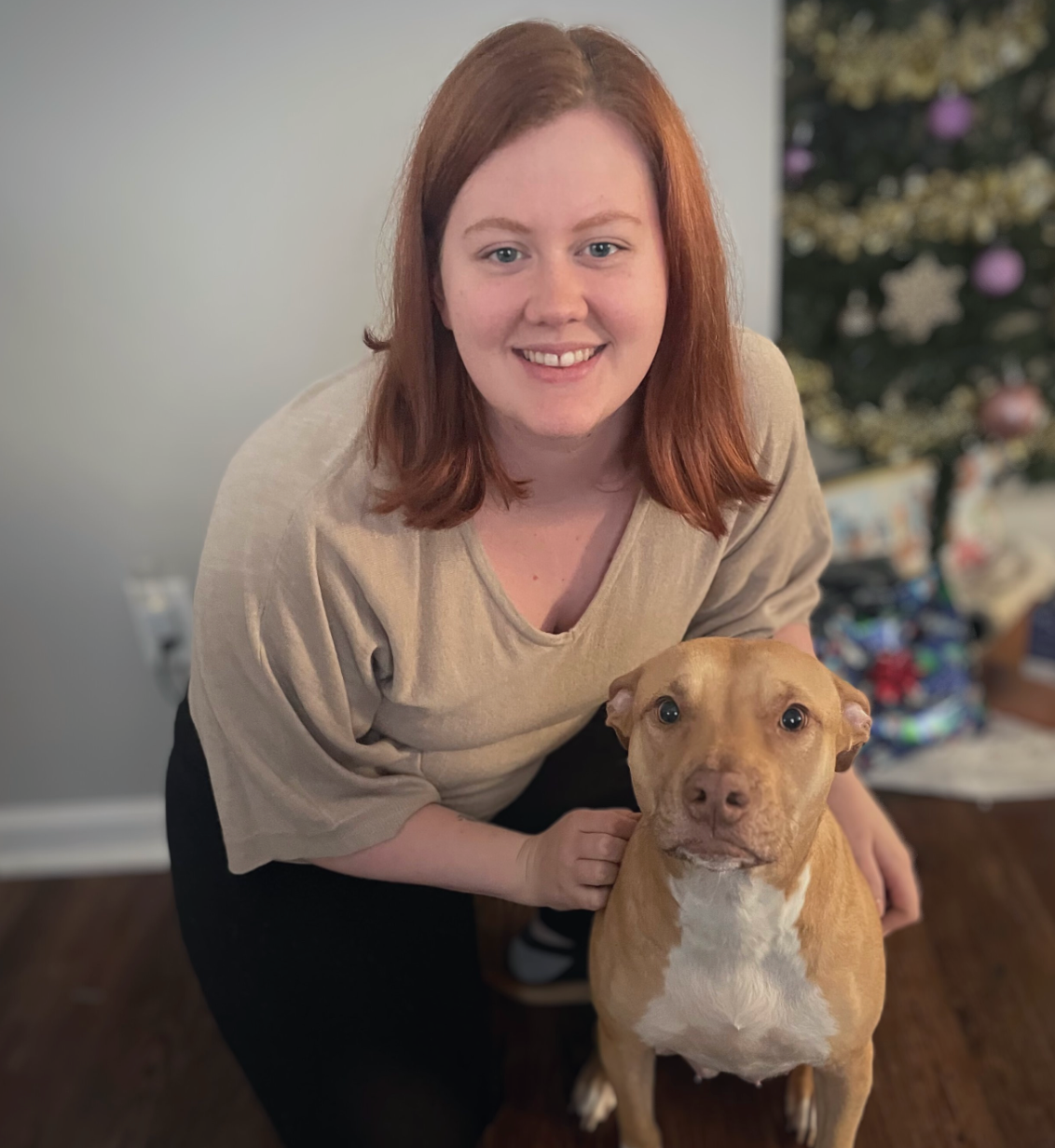 Smiling woman crouches beside a tan dog indoors, with a blurred decorated tree in the background