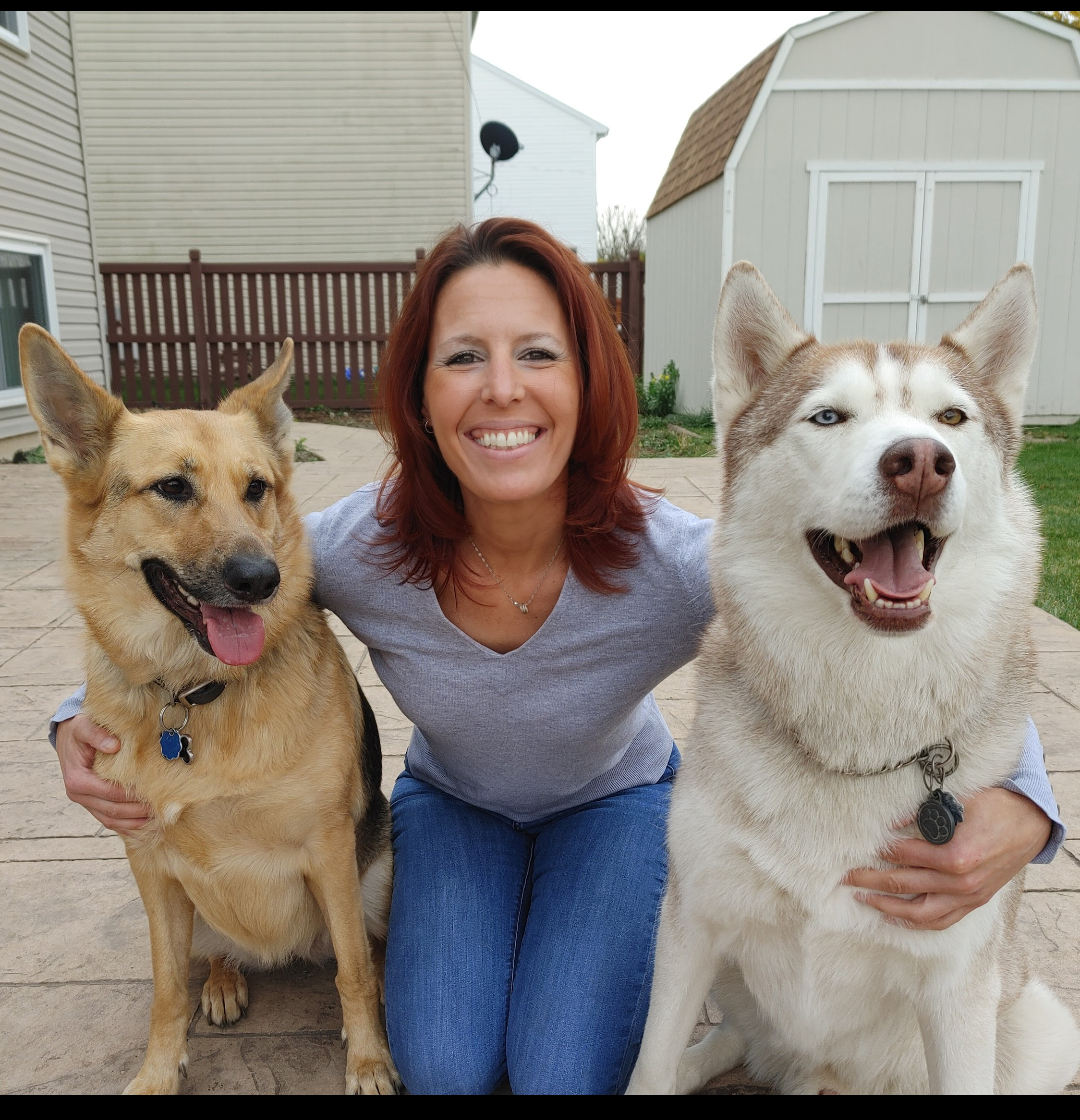 Woman kneeling between two happy dogs in a backyard, one tan and one white, both panting with tongues out