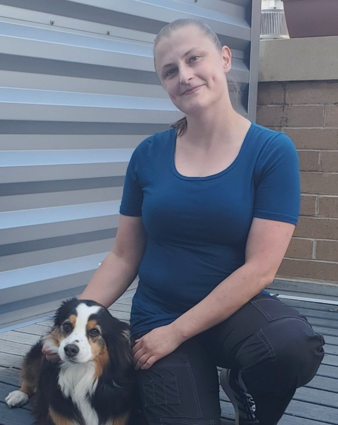 Woman crouching beside a black-and-white dog outdoors near a corrugated wall and brick steps
