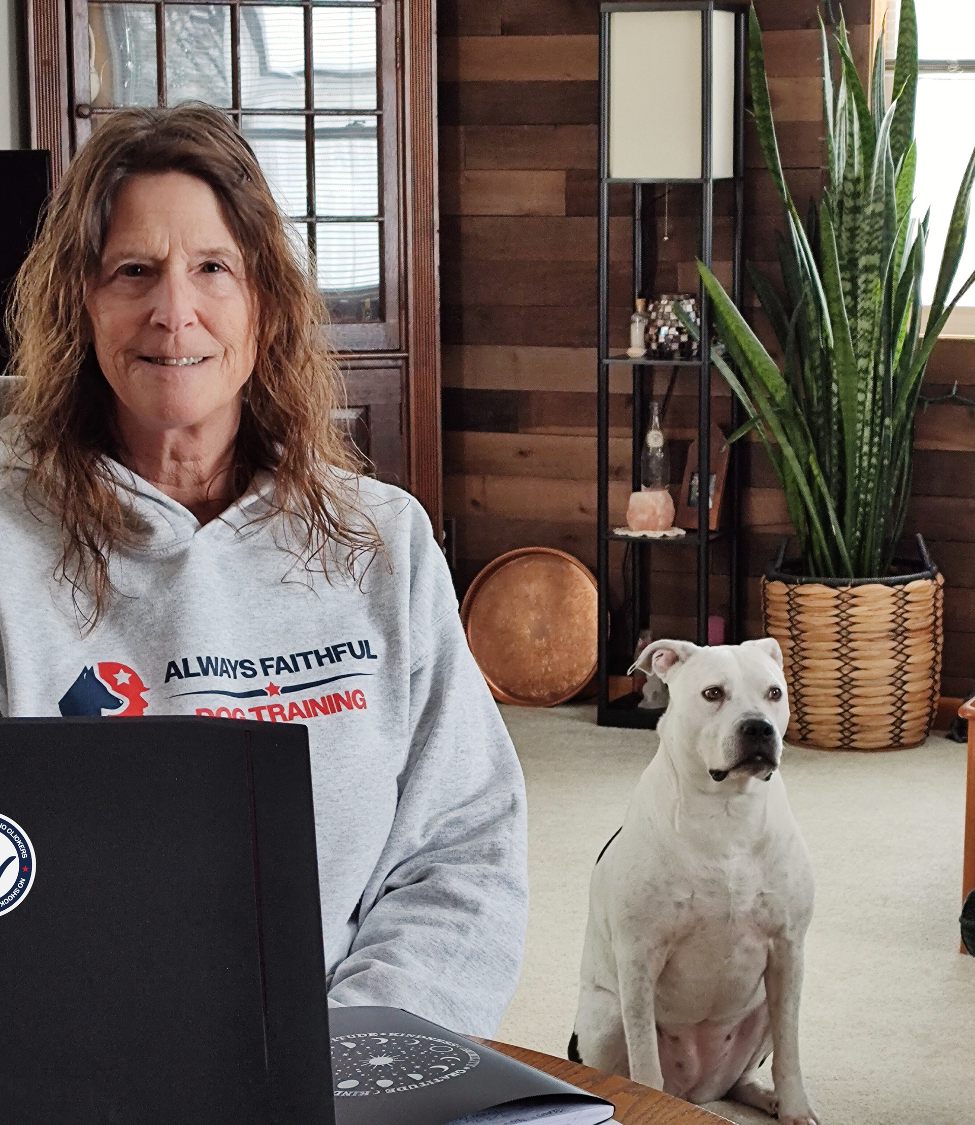 Woman seated indoors beside a white dog, with a laptop in front of her.