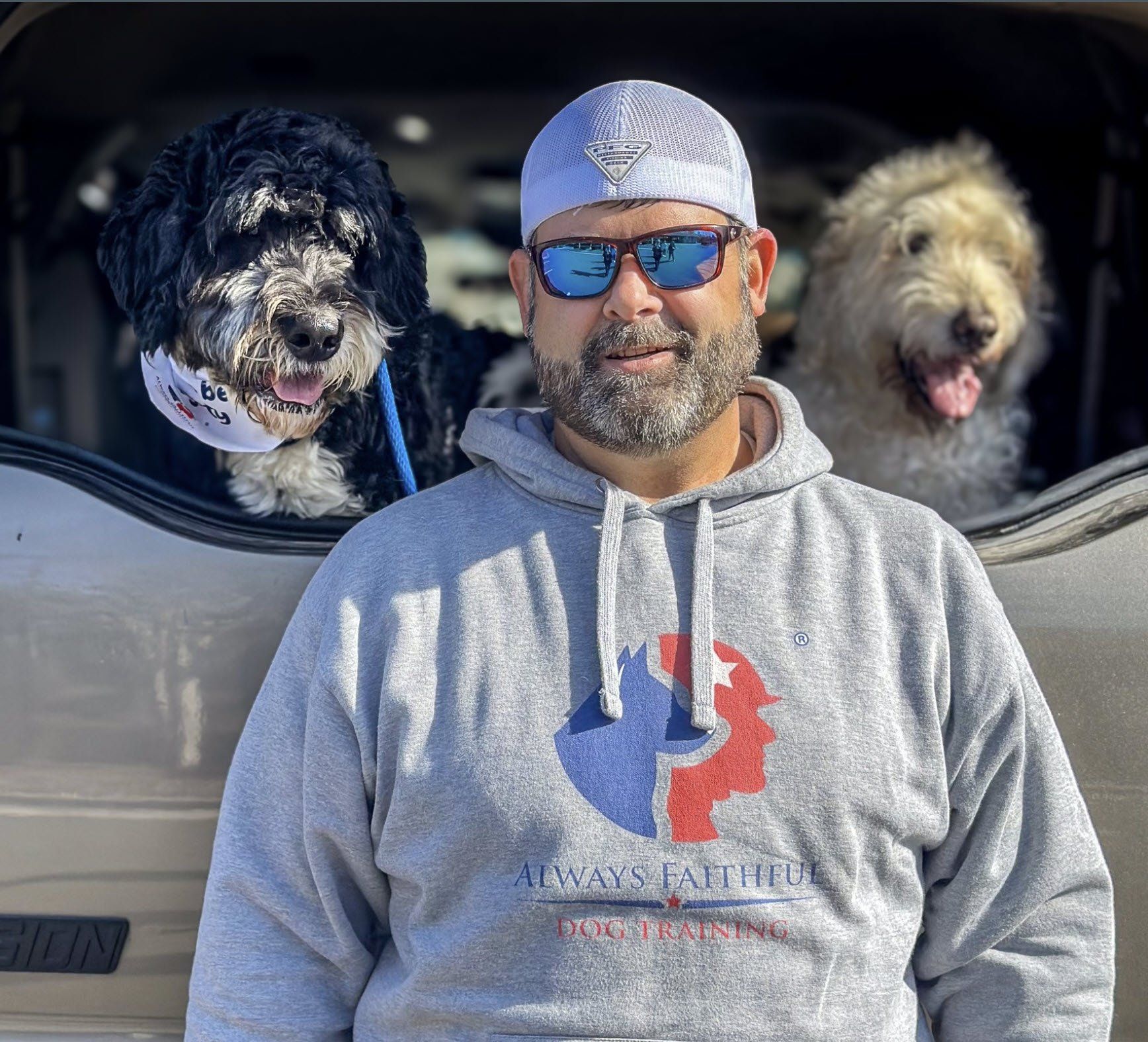 Man in gray hoodie and cap standing with two dogs peeking from a car window