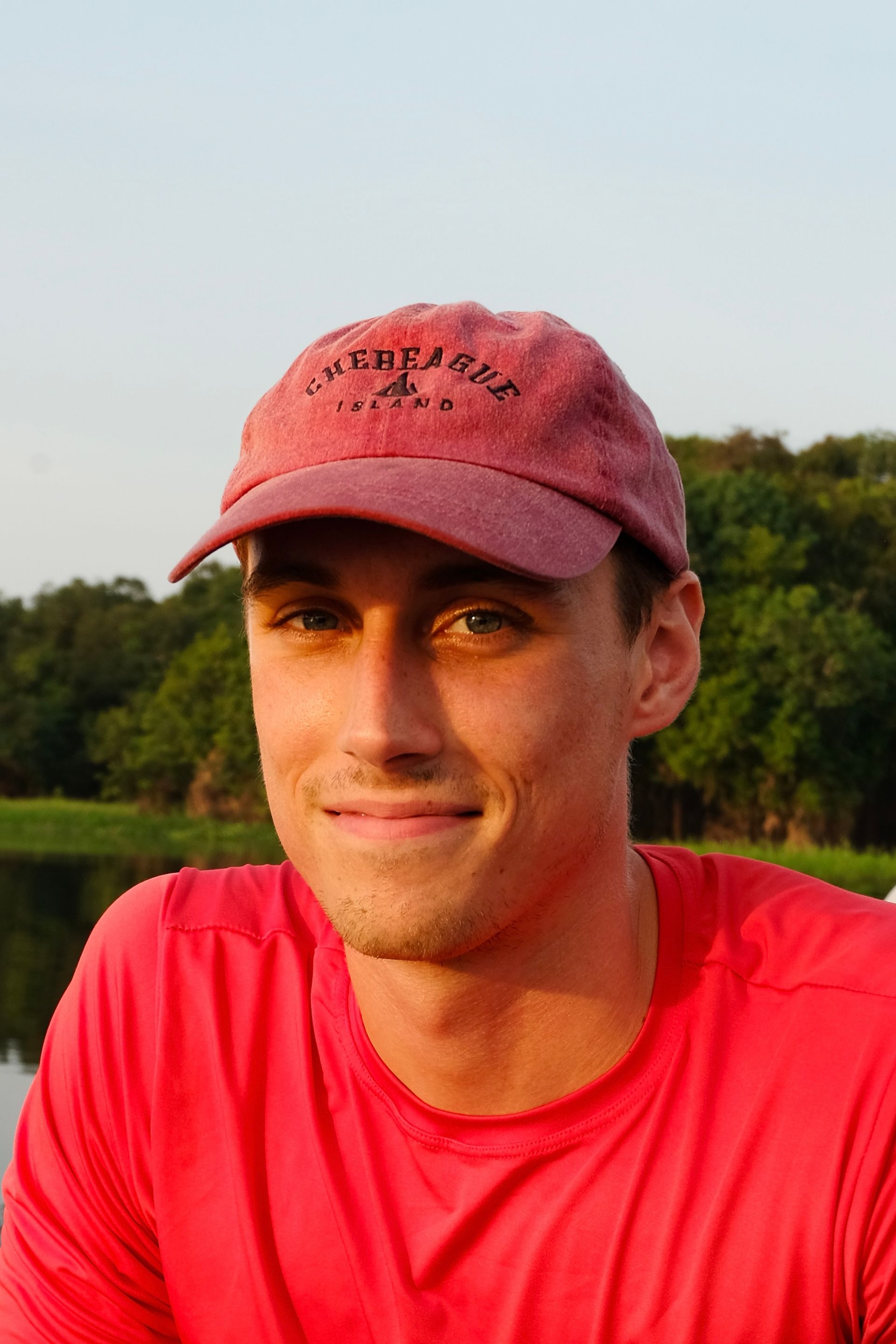 Man in red shirt and maroon cap outdoors, smiling with trees and sky behind him
