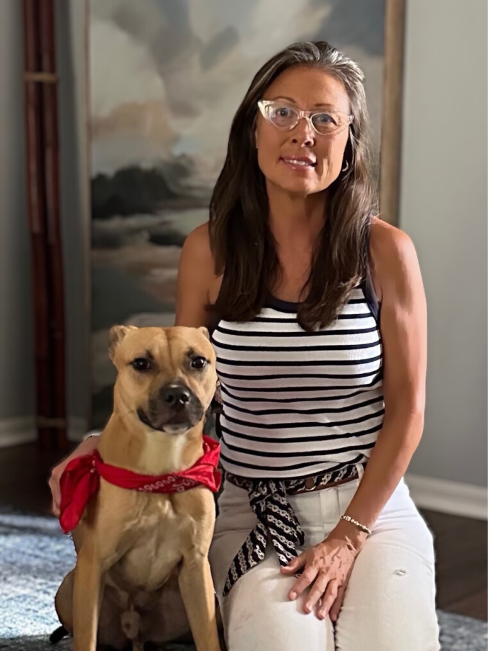 Woman in striped tank top with tan dog wearing a red bandana, sitting indoors.