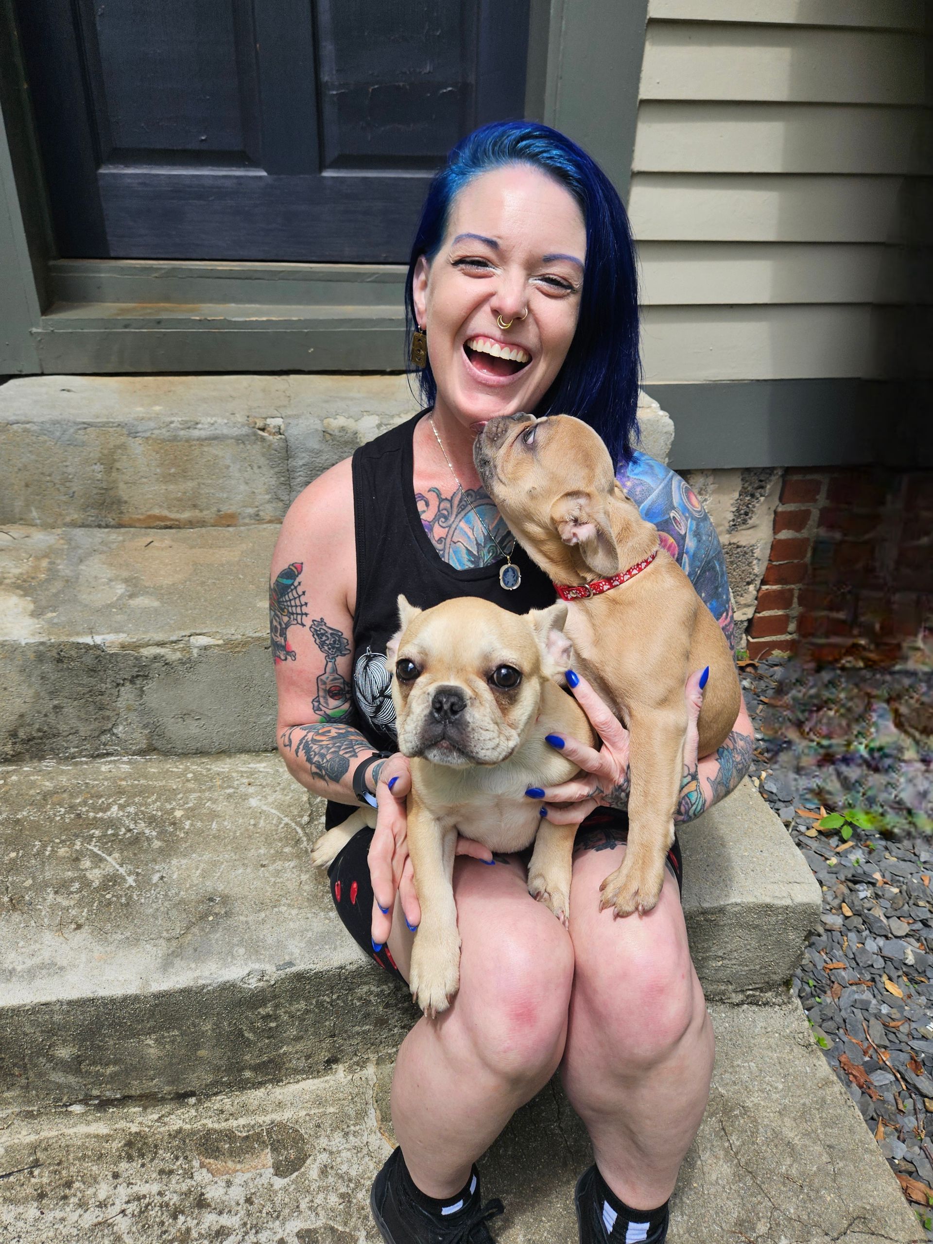 Smiling person with blue hair holding two small dogs on outdoor steps