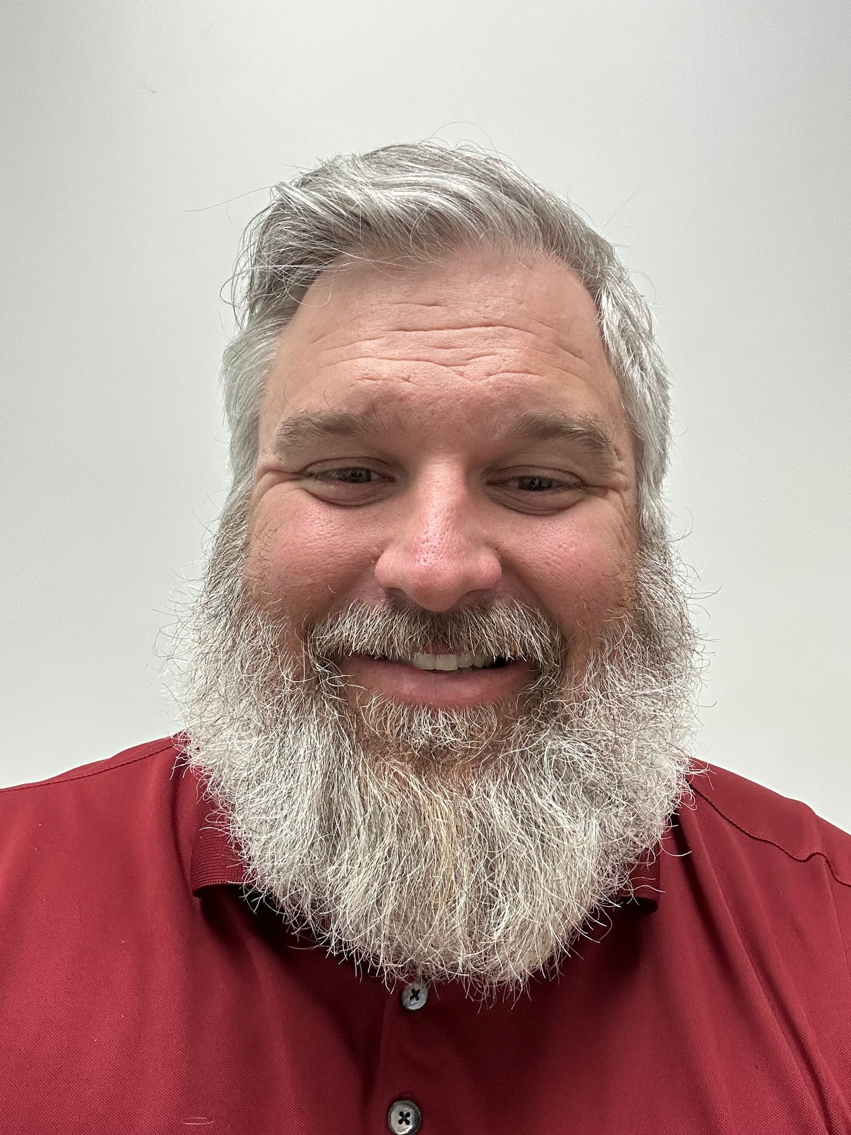 Smiling man with a gray beard wearing a red shirt against a plain light background