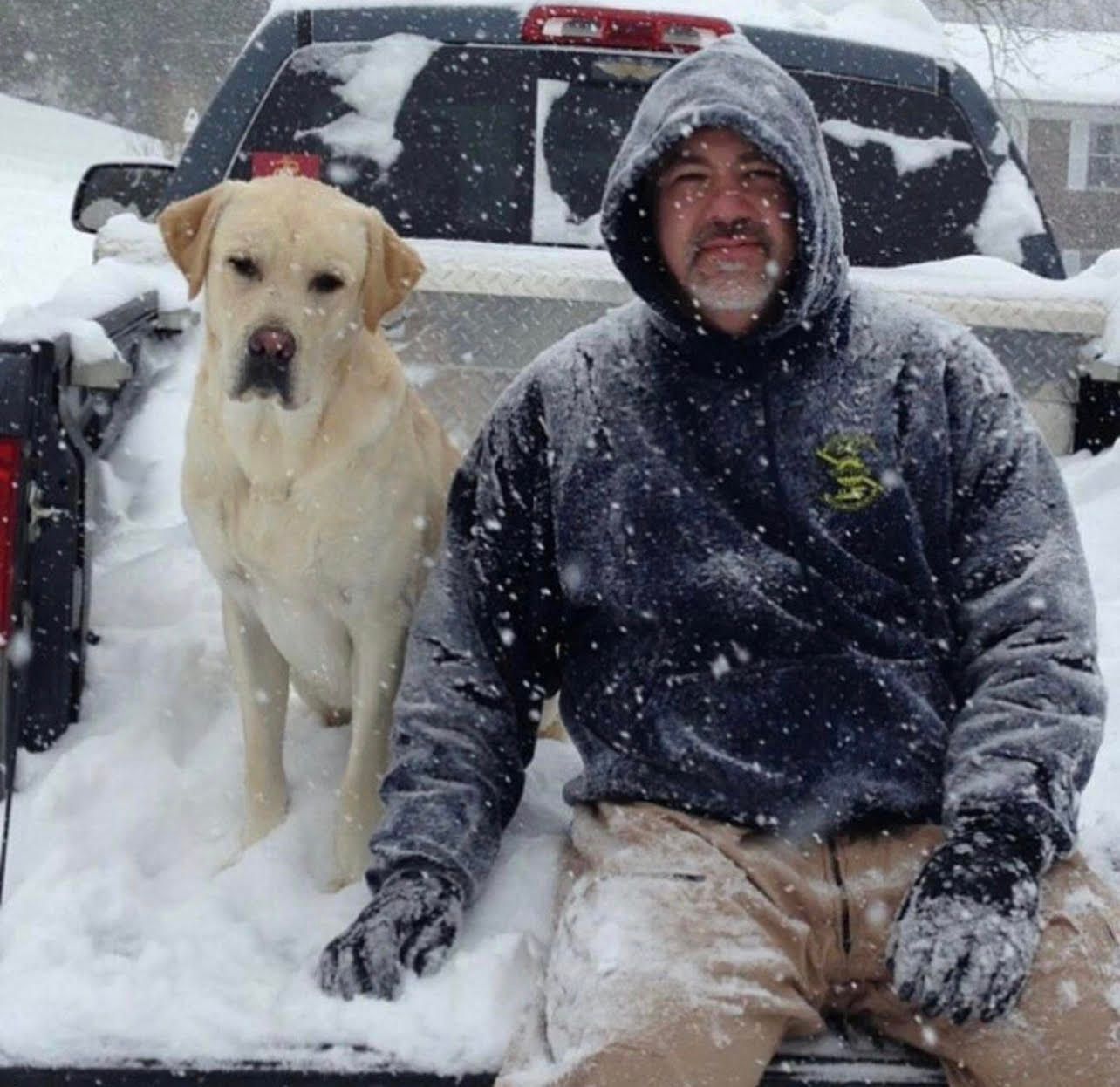 Man and dog sitting in a snow-covered truck bed during heavy snowfall