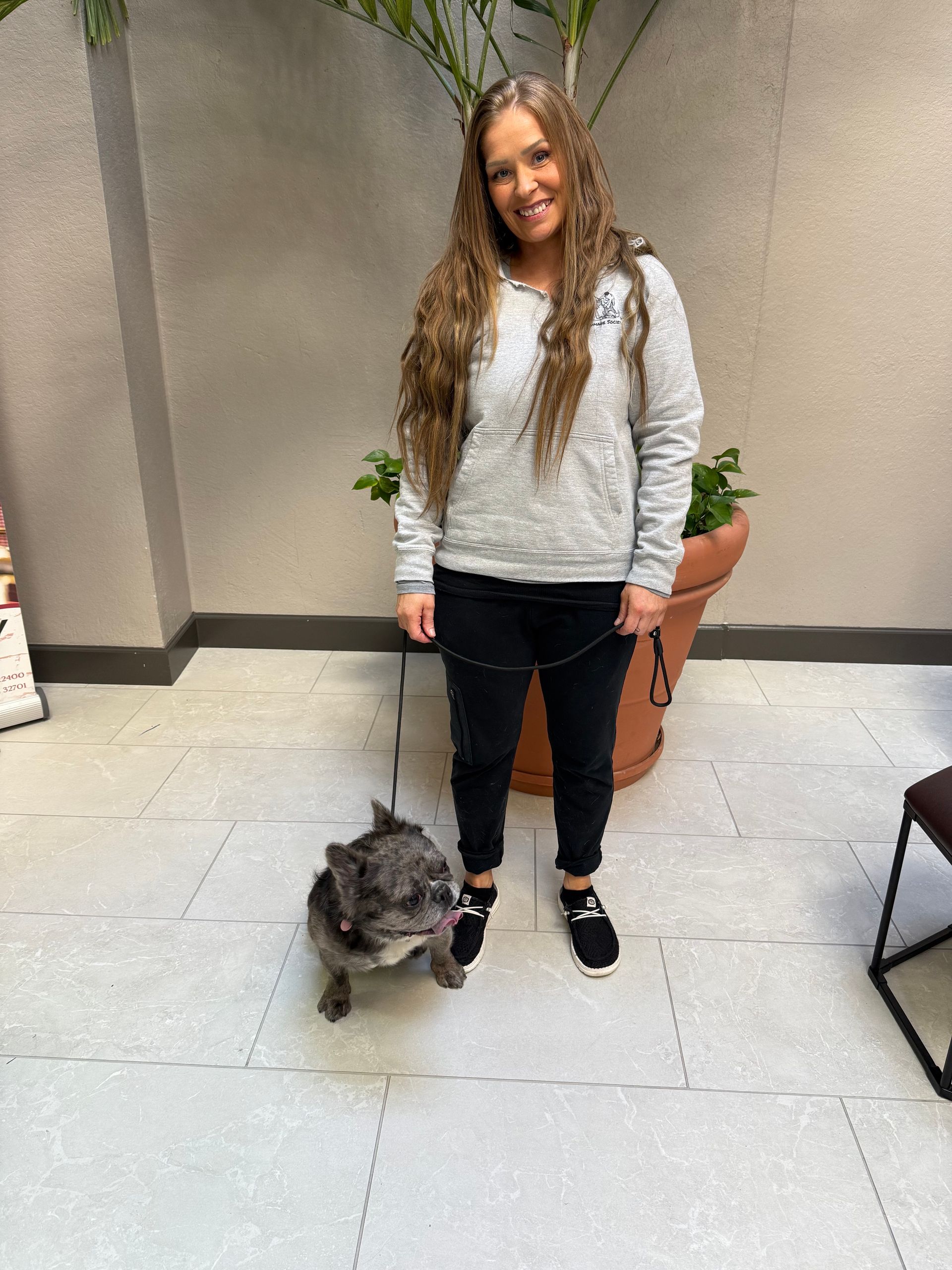 Woman standing with a small dog on a patio beside potted plants, wearing a gray sweater and black pants.