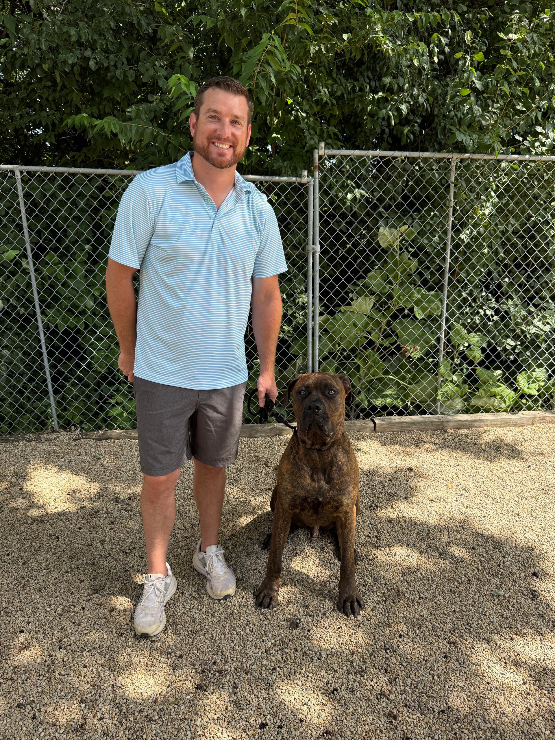 Man standing with a brown dog in a gravel yard by a chain-link fence and trees