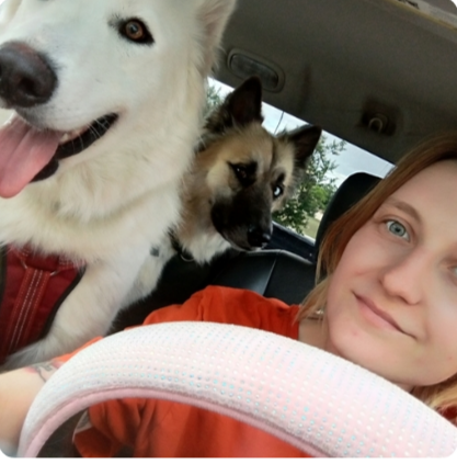 Woman taking a selfie in a car with two dogs, one white and one tan, both leaning into the frame