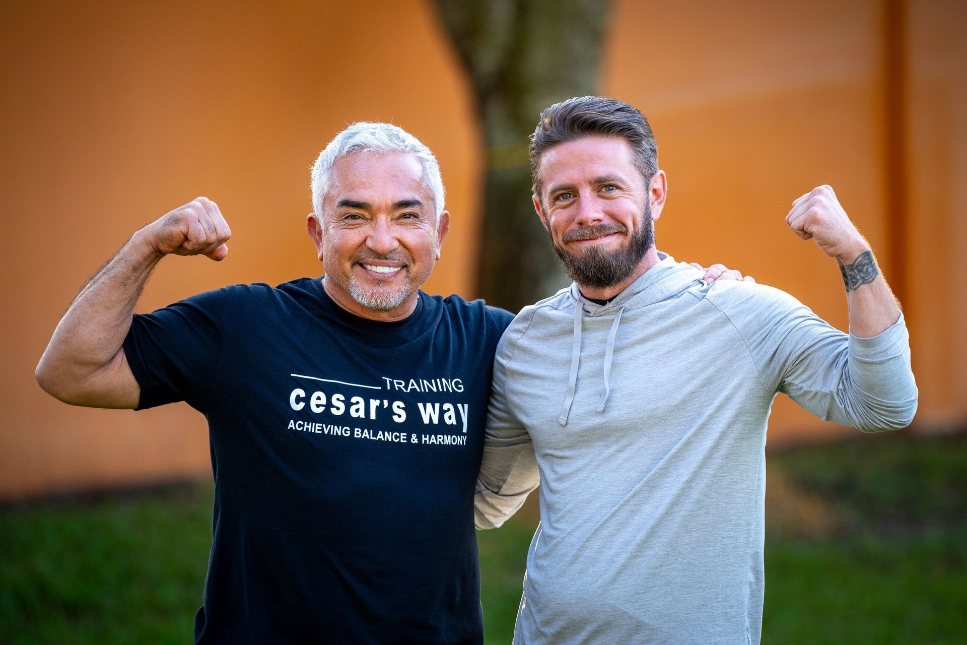 Two smiling people flexing their arms outdoors in front of an orange wall