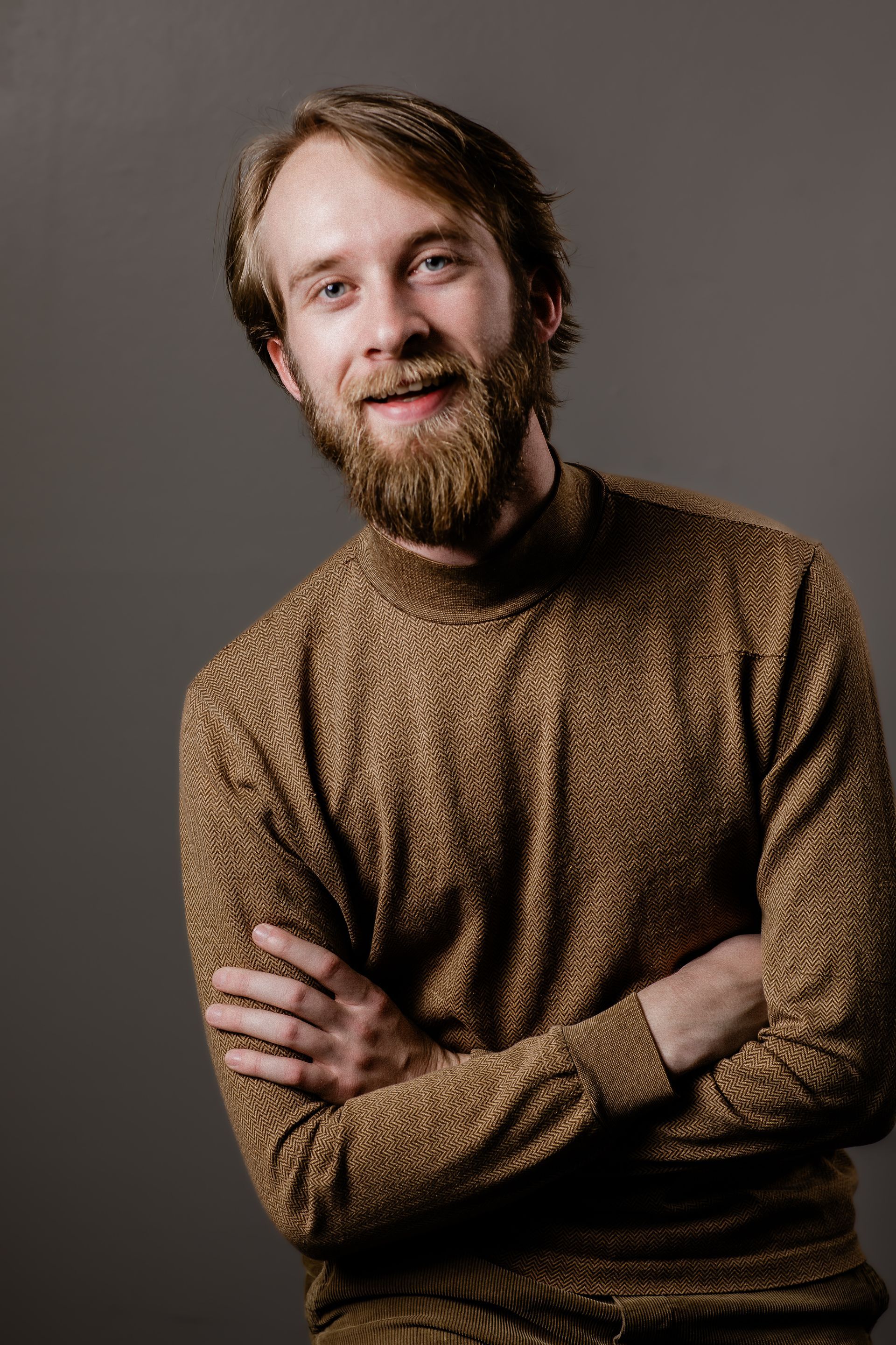 Smiling bearded person in a brown sweater with arms crossed against a plain gray background