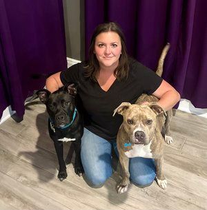 Woman kneels with two dogs in front of purple curtains, one black and one tan-gray, both wearing collars.